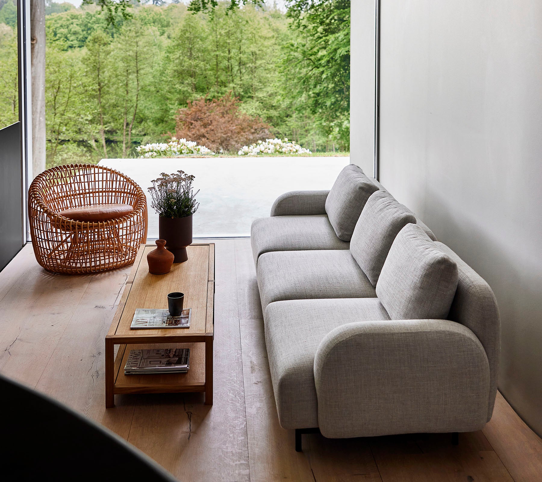 Modern soffa i ljus tyg i kombination med ett träkaffebord och en korgstol, omgiven av grönska. Naturligt solljus förbättrar den lugna atmosfären.