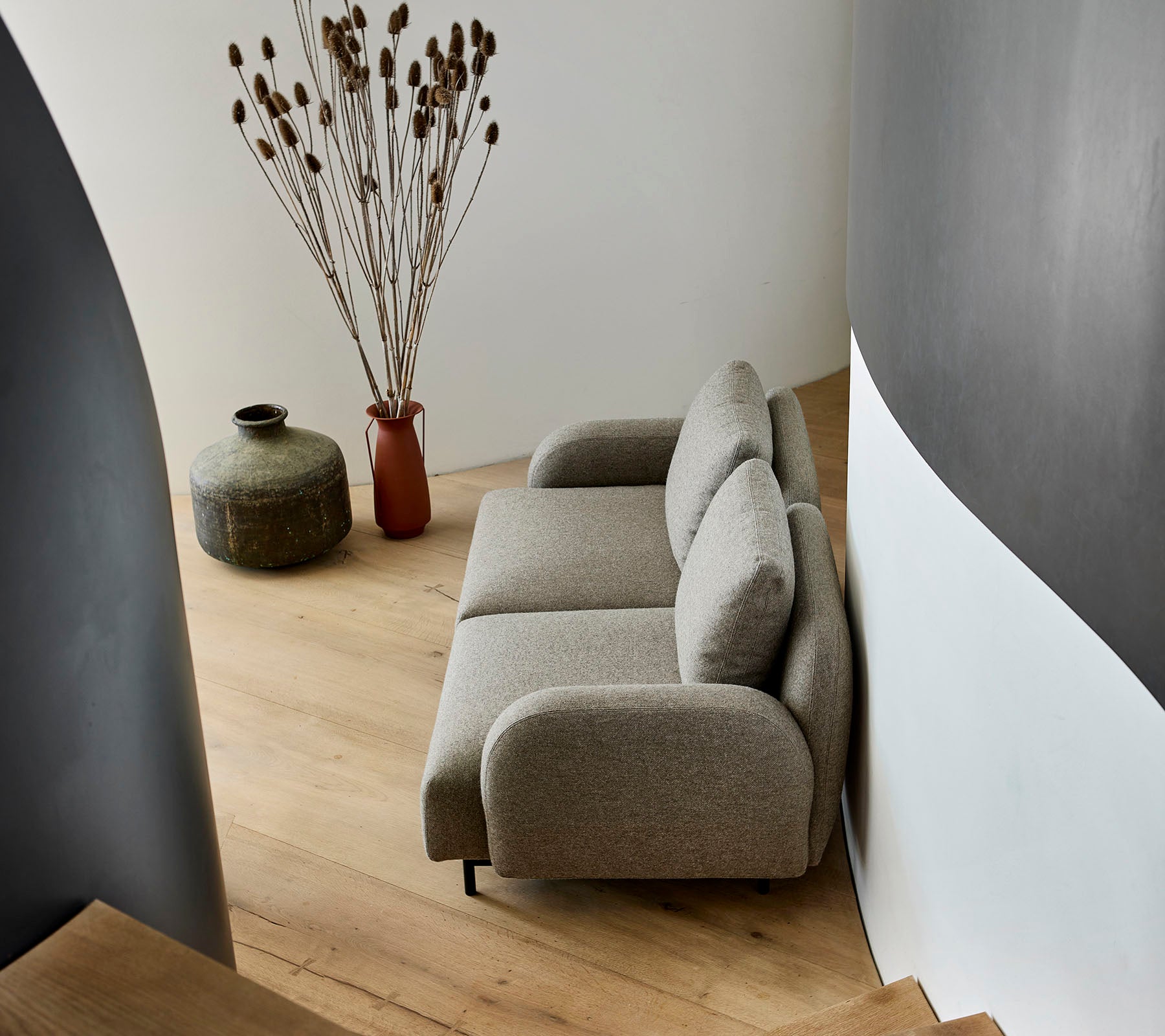 En mysig beige soffa placerad bredvid en dekorativ vas och torra växter, mot en böjd vägg och trägolv.