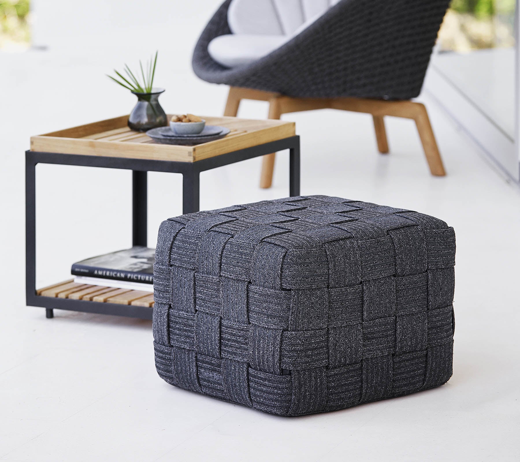 En texturerad grå vävd pouf placerad nära ett träsidebord med minimalistisk design, som har små dekorationsföremål.