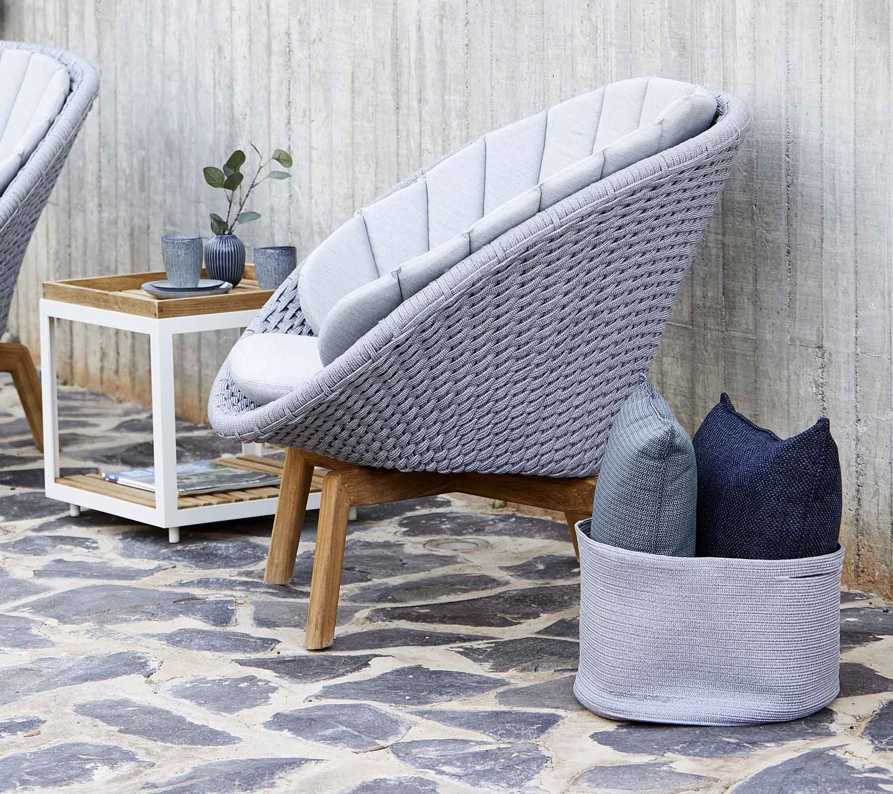 Modern grå loungestol med en vävd design, kompletterad med kuddar och ett litet träsoffbord. Stengolvet förbättrar estetiken.