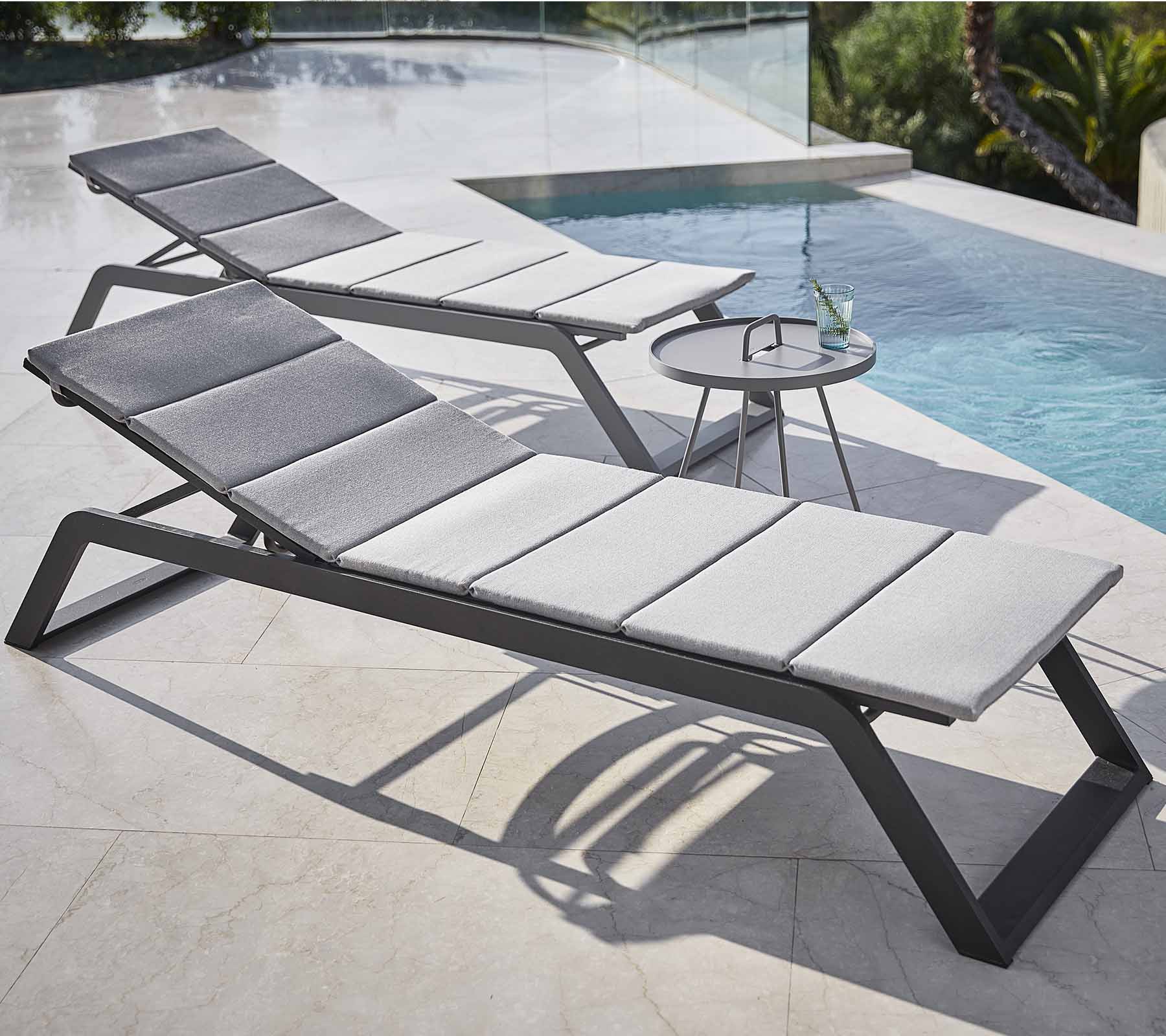 Två stiliga loungestolar med grå kuddar är placerade vid en pool, tillsammans med ett litet sidobord med en drink.