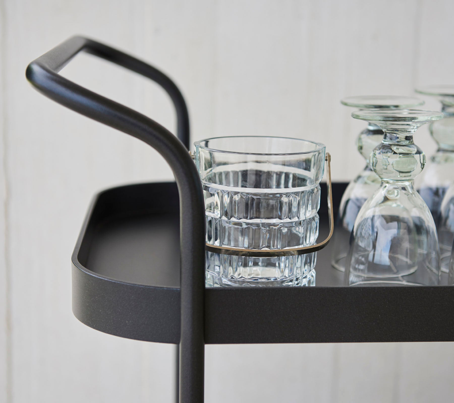 Stilren serveringsvagn med ett smalt handtag, som har ett glas tumbler och flera klara glasföremål på en plan yta.