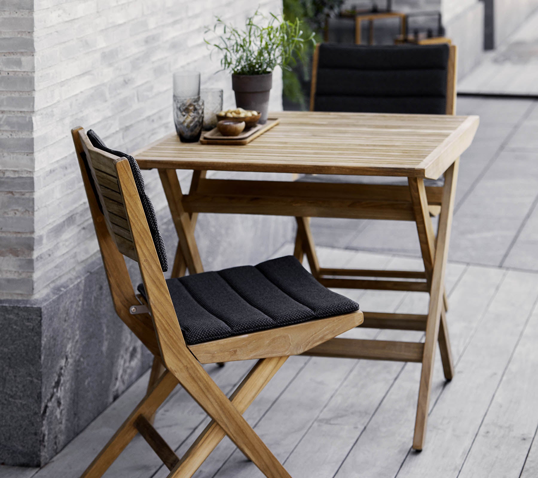 Träbord- och stolset med svarta kuddar, prytt med krukväxter och dekorativt glas. Perfekt för en mysig miljö.