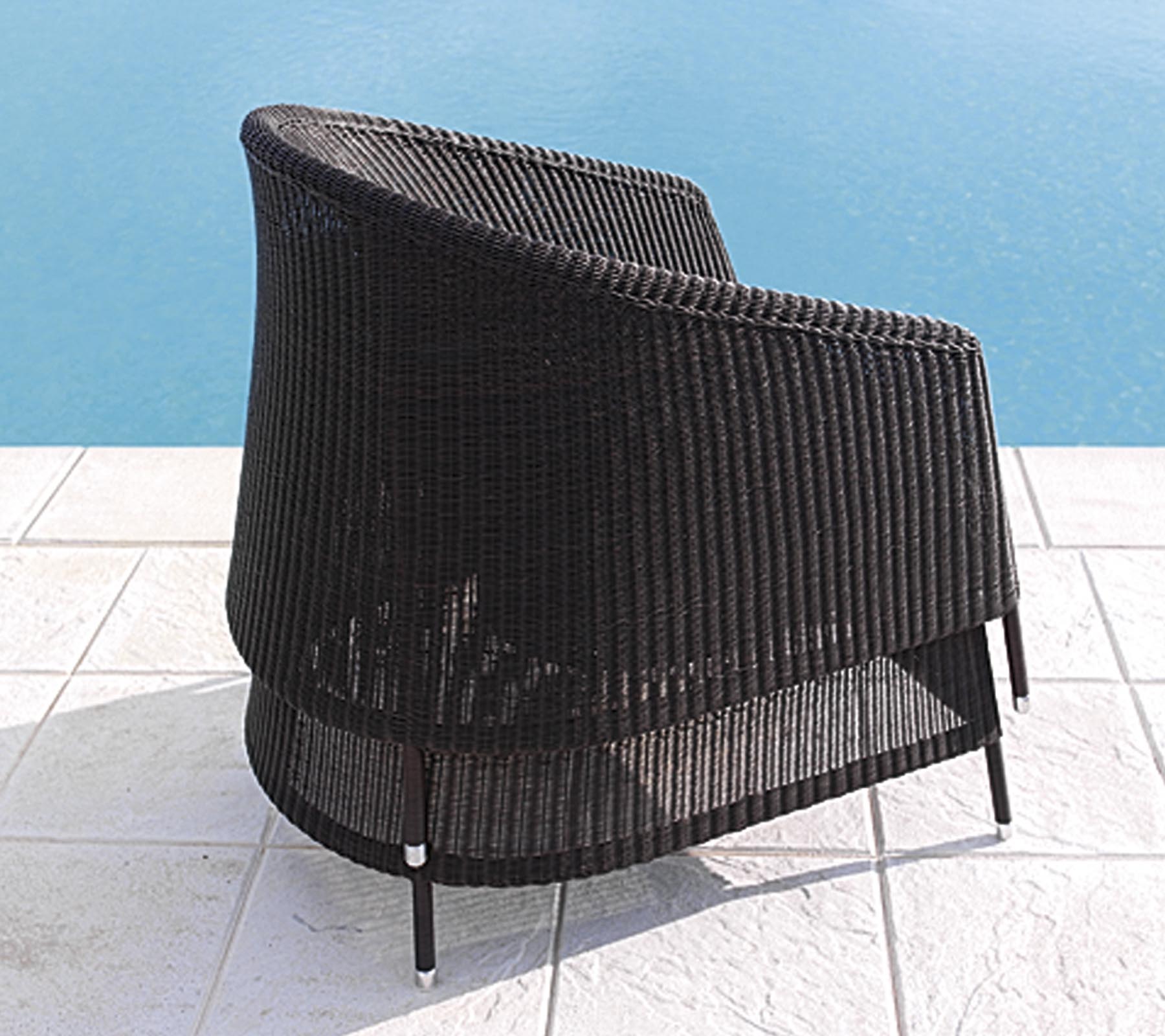 Kurvad svart rottingsstol med en elegant design, placerad nära en pool på vit kakelgolv.