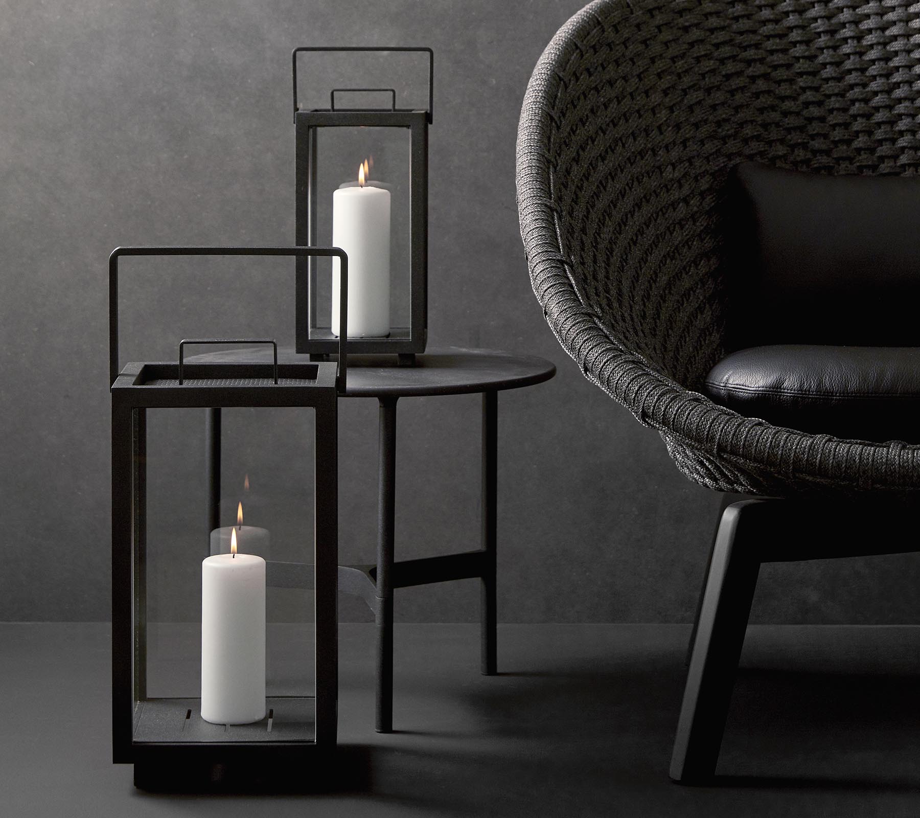 Två stiliga ljuslyktor med vita ljus, åtföljda av en modern, texturerad stol på ett minimalistiskt bord mot en mörk bakgrund.