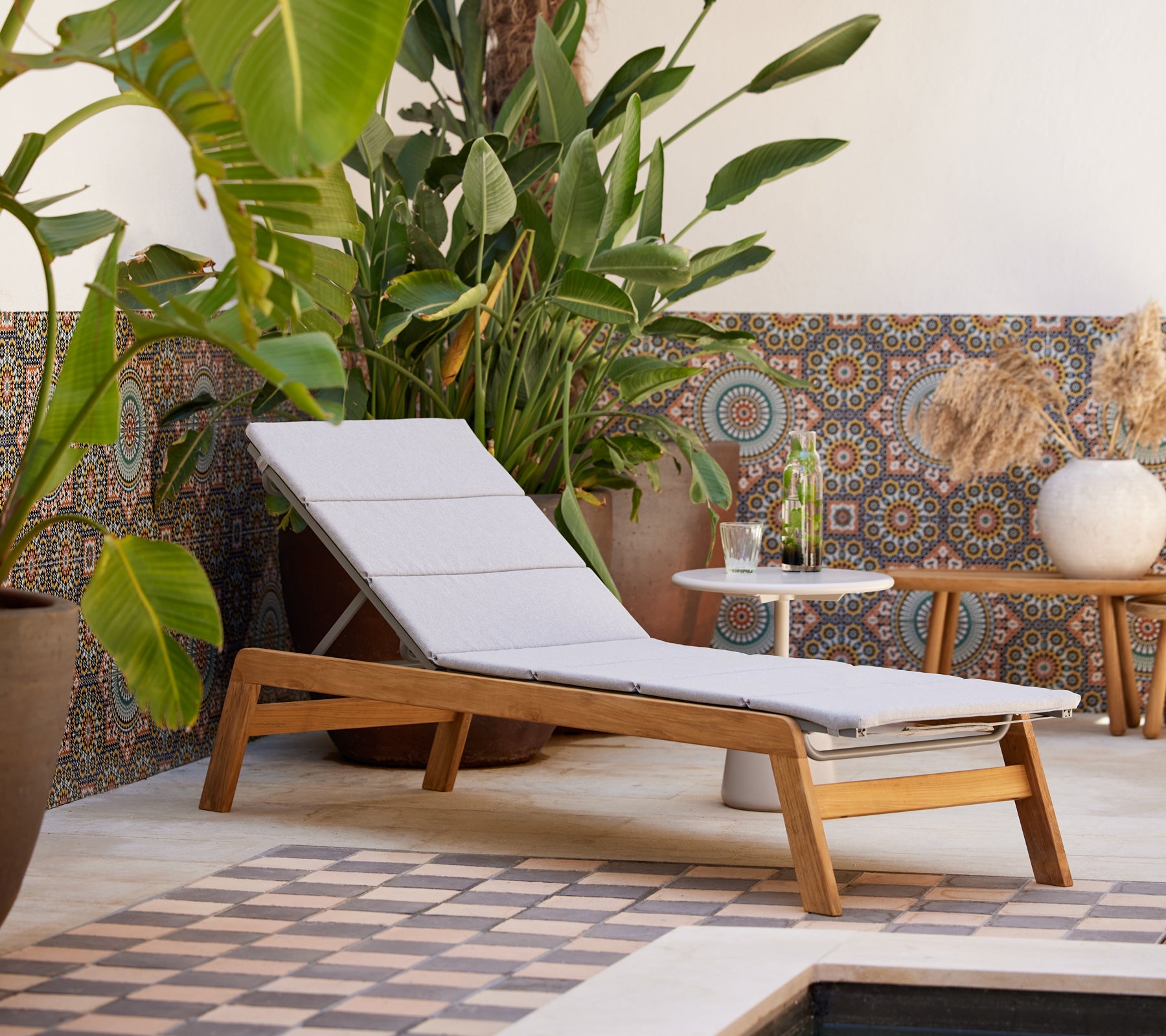 En modern loungestol med ett ljust kudde, ihop med ett sidobord, omgiven av frodig grönska och dekorativt kakel i bakgrunden.