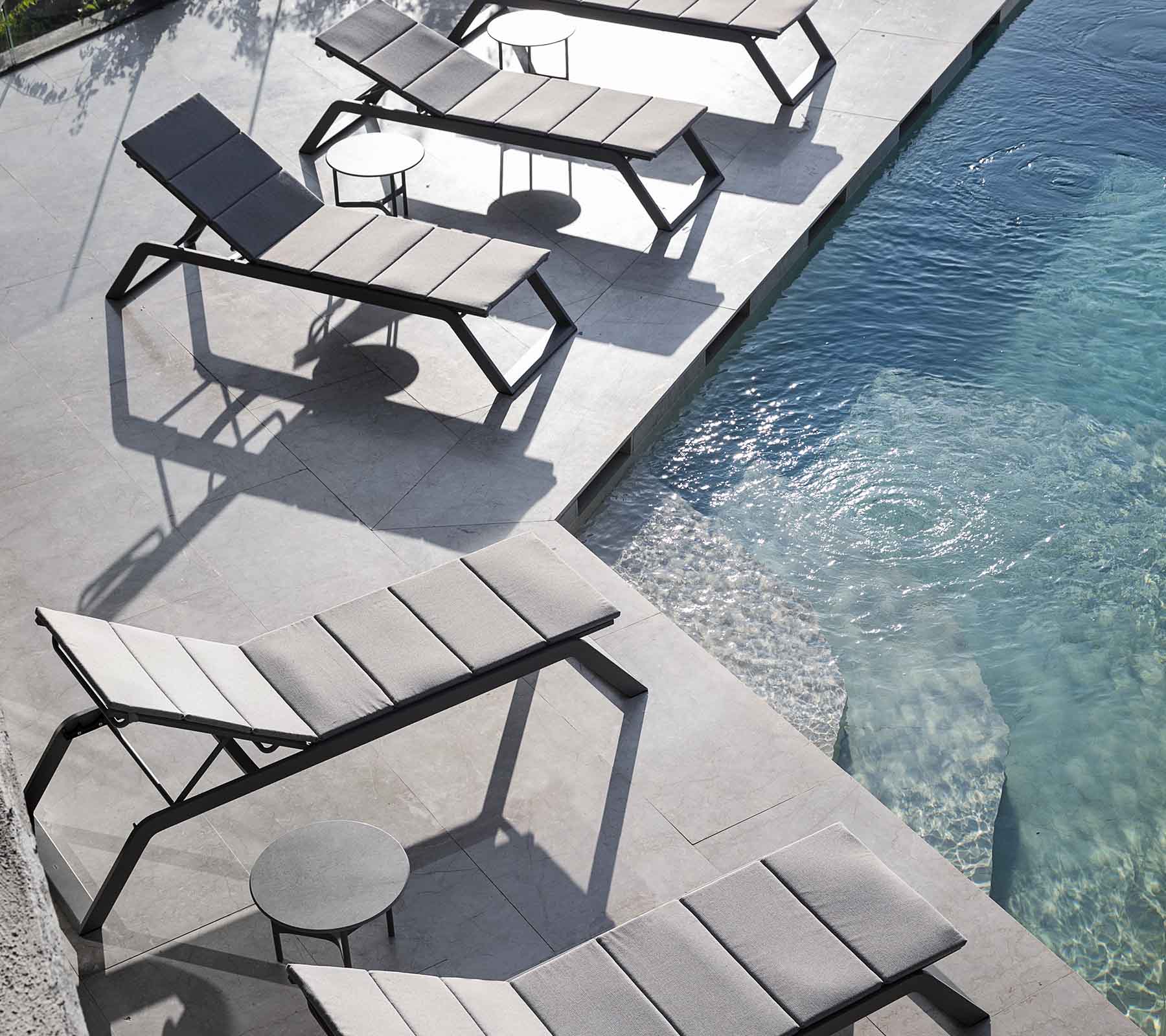 En rad av moderna loungefåtöljer med ett litet sidobord vid en swimmingpool, som kastar skuggor på pavéet.