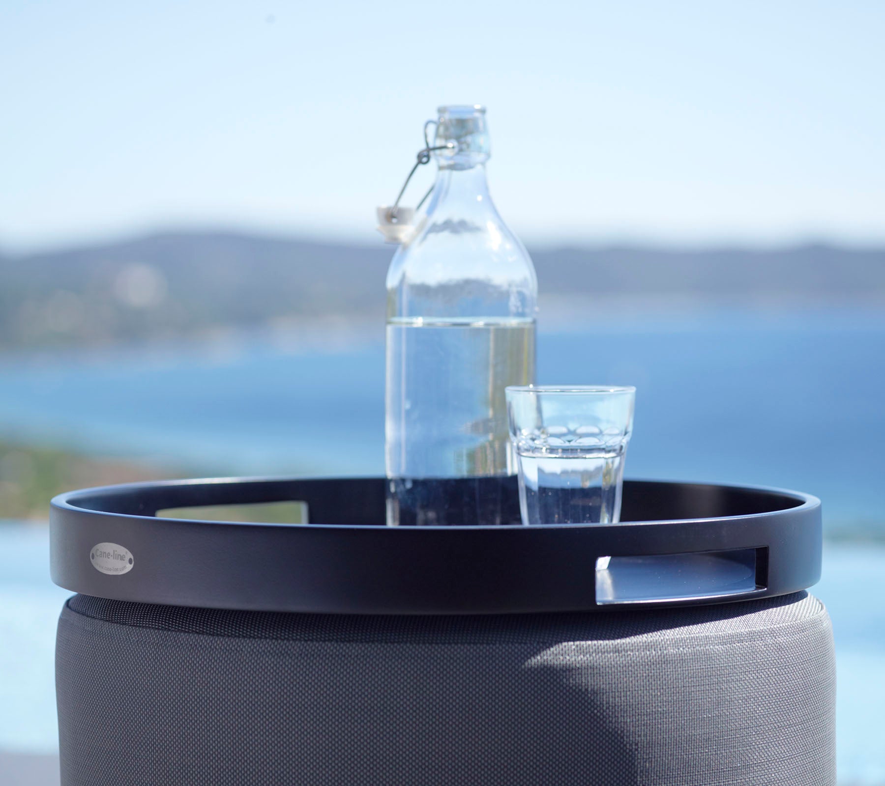 En svart rund bricka håller ett glas vatten och en glasflaska, mot en naturskön bakgrund av havet och avlägsna kullar.