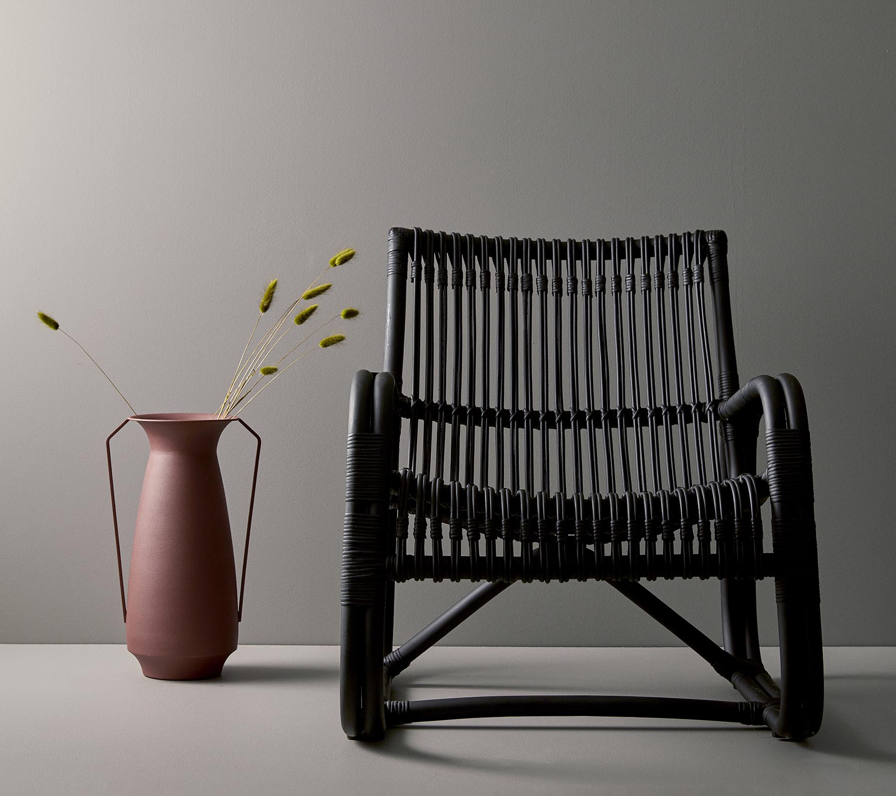 Stilfull svart vävd stol bredvid en rosa vas med gröna stjälkar, mot en minimalistisk grå bakgrund.