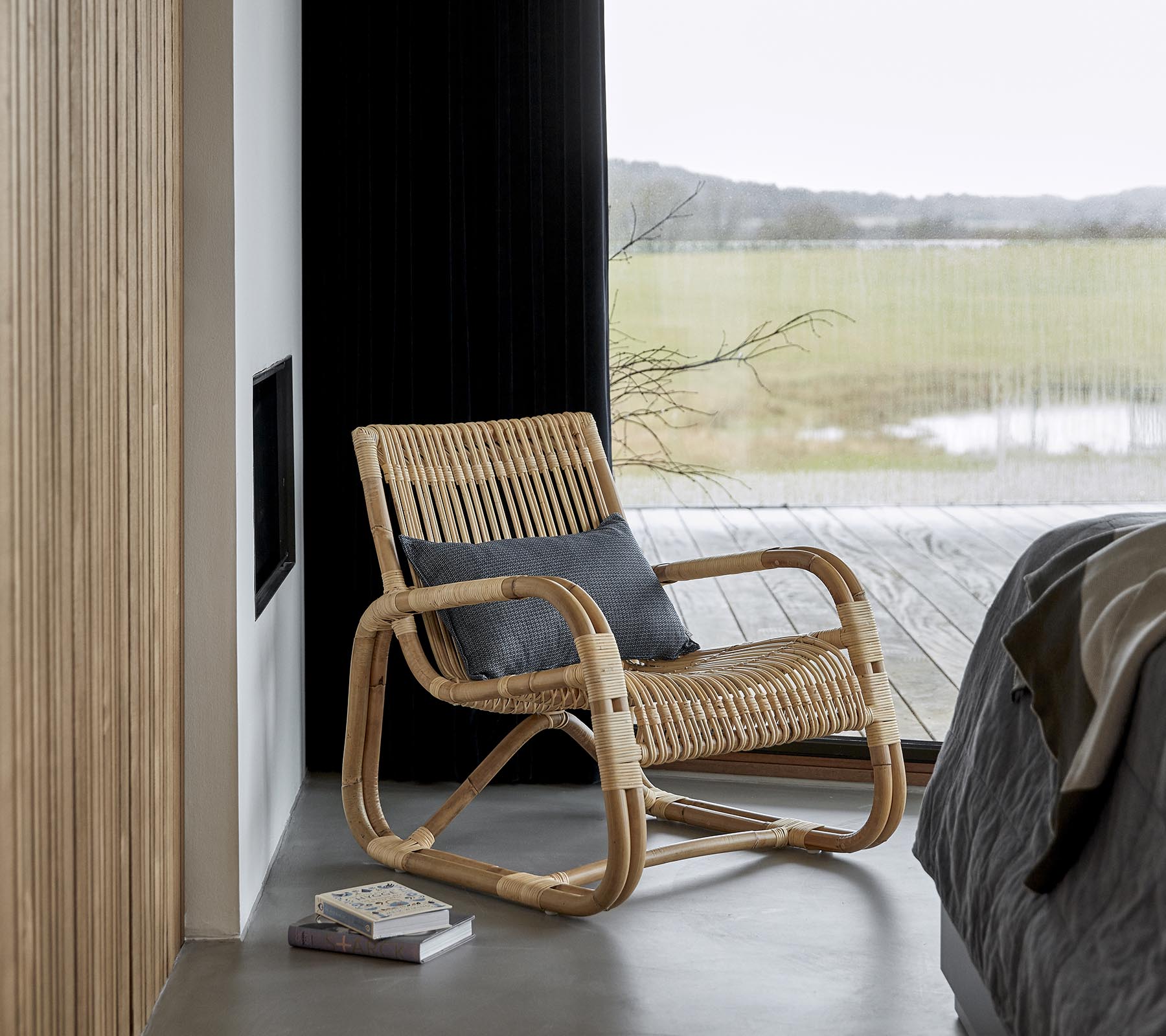 En stilren gungstol gjord av naturliga material, placerad intill en säng och en bok, med en naturskön utsikt synlig genom stora fönster.