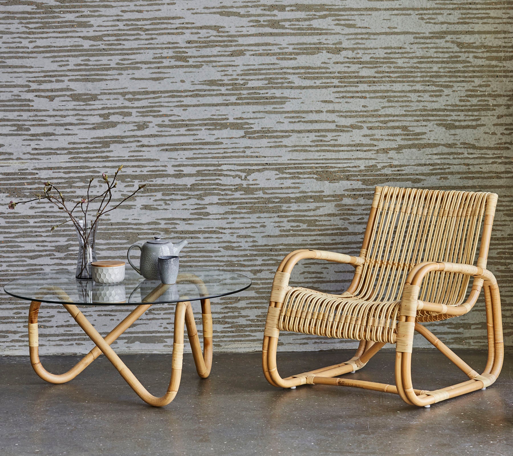 Rattanstol och bord med glasskiva med dekorativa föremål, placerade mot en texturerad vägg. En minimalistisk och modern inredningsstil.