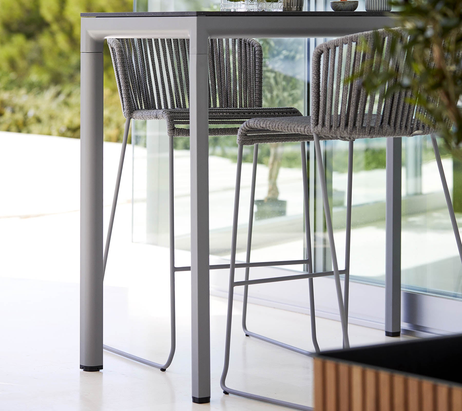 Ett elegant, modernt bord med matchande höga stolar som har en minimalistisk design och en stilren metallram, kompletterat med naturlig grönska.