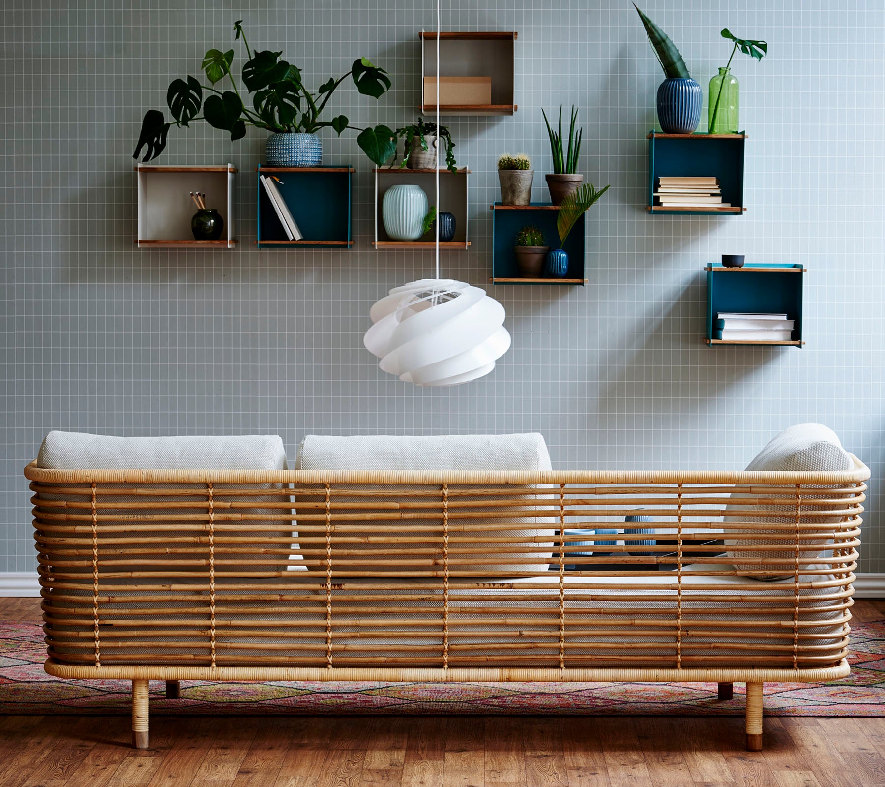En stilren rattan-soffa med ljusa kuddar mot en strukturerad vägg, kompletterad med väggmonterade hyllor som visar växter och dekorativa föremål.