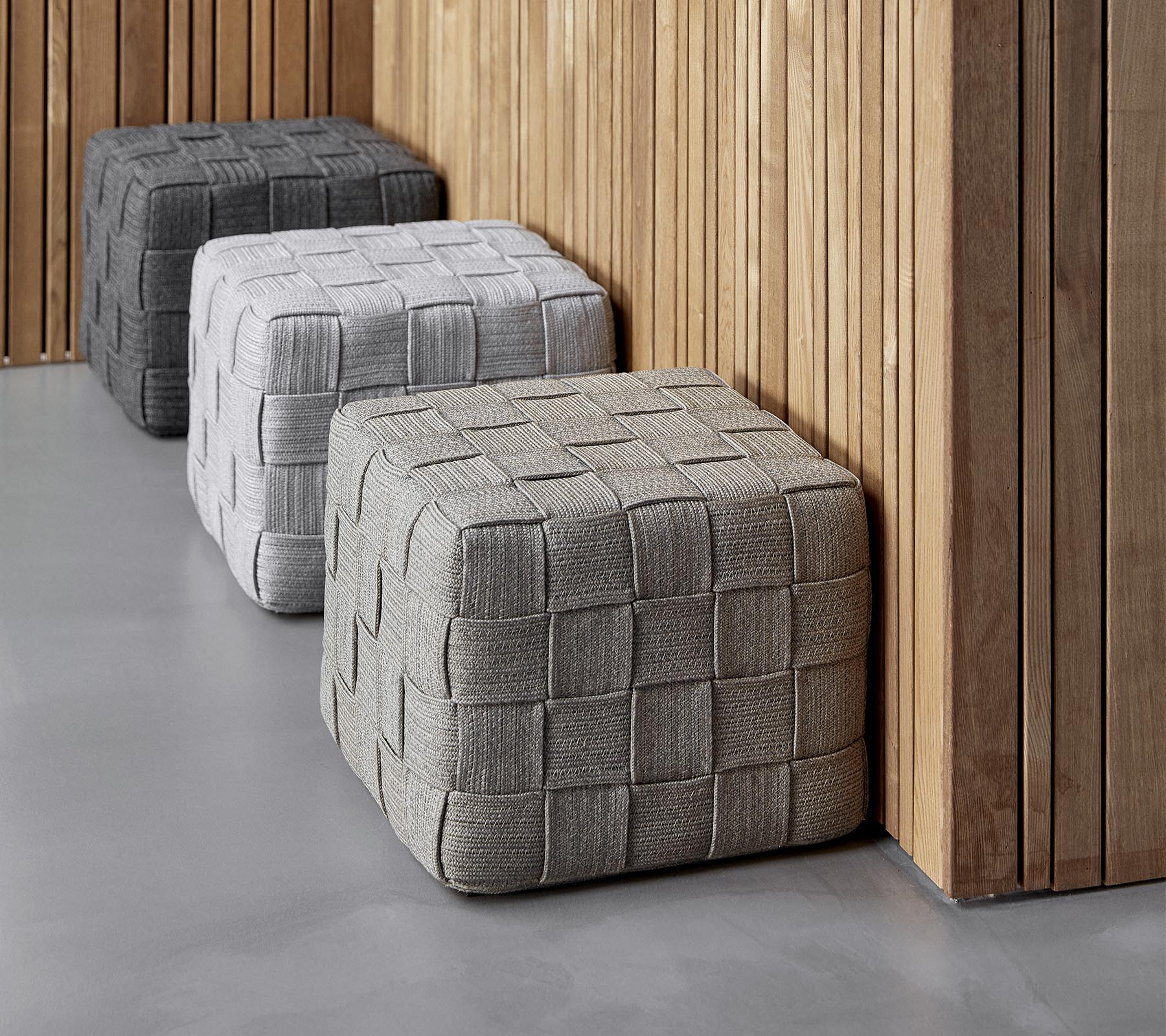 Tre texturerade, fyrkantiga ottomaner i olika nyanser av grått är placerade mot en trävägg, vilket ger en modern känsla till miljön.
