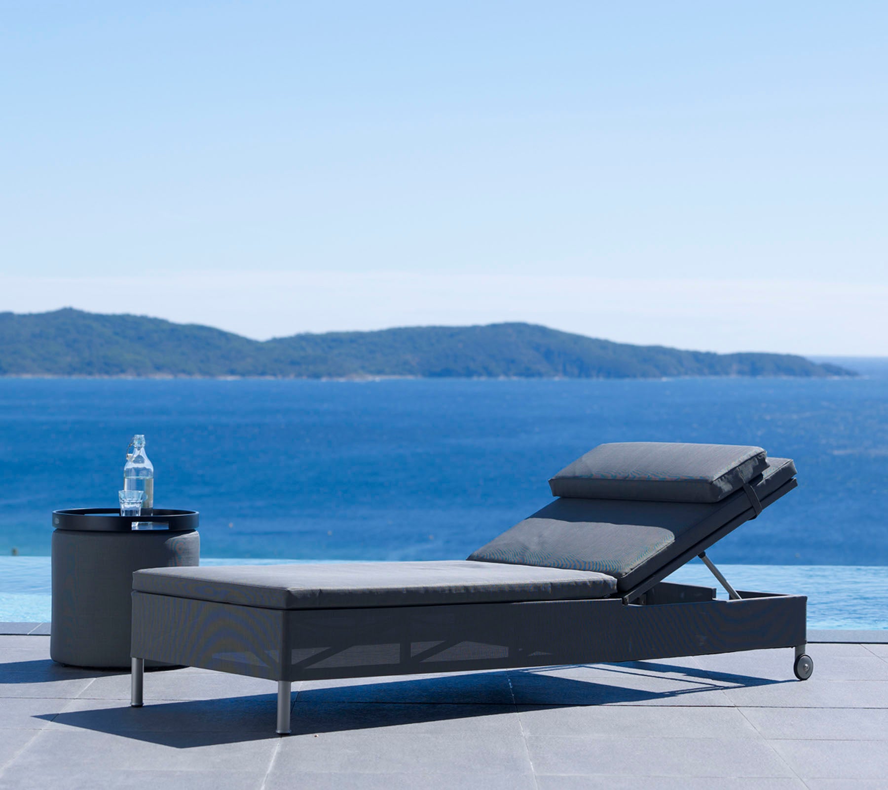En modern loungestol och sidobord vid ett stilla vattendrag med avlägsna högar under en klar himmel, perfekt för avkoppling.