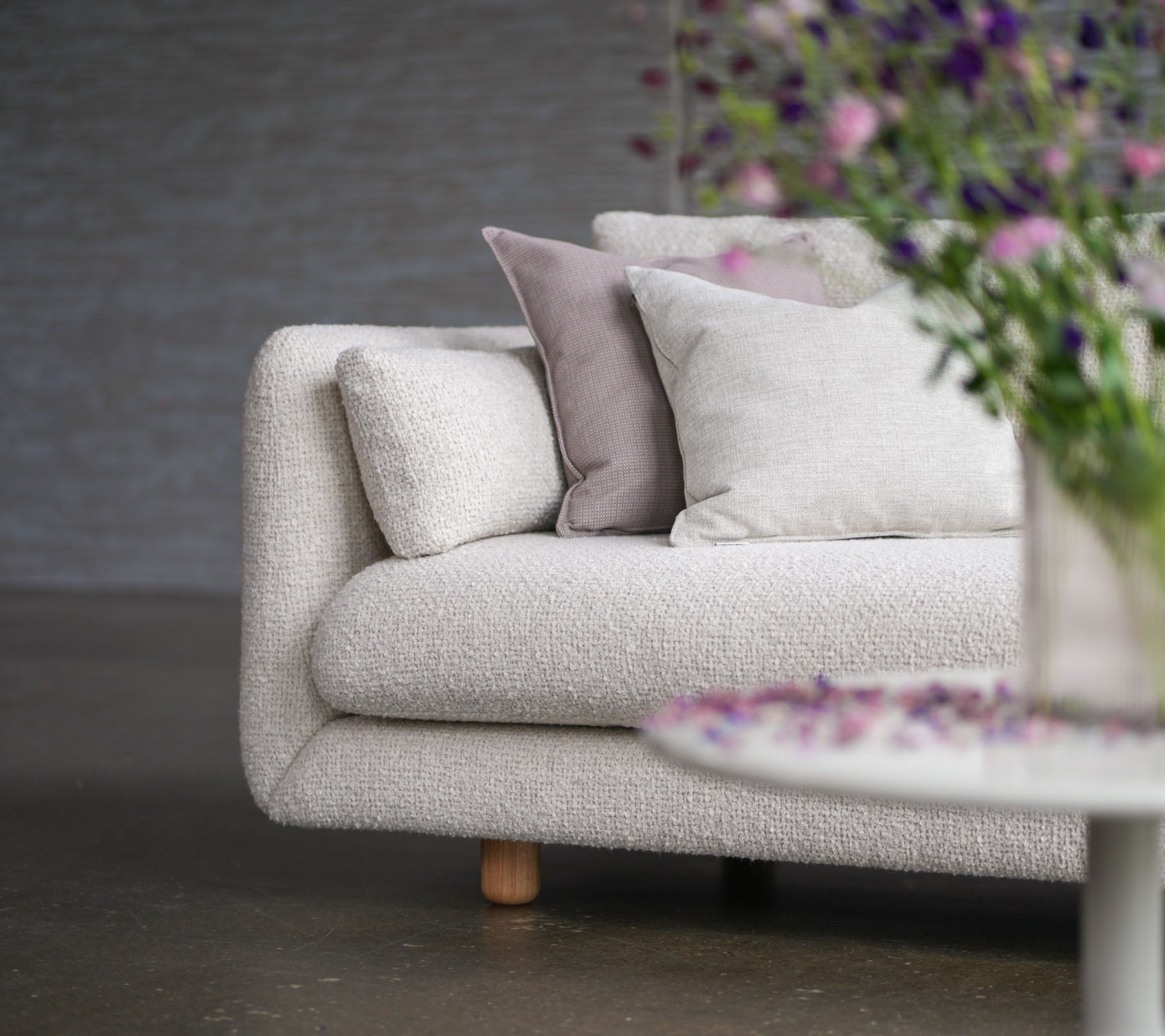 Mjuk soffa med sandfärgad klädsel, dekorativa kuddar, teakben och en vas med blommor som visar Cane-lines stil och elegans.