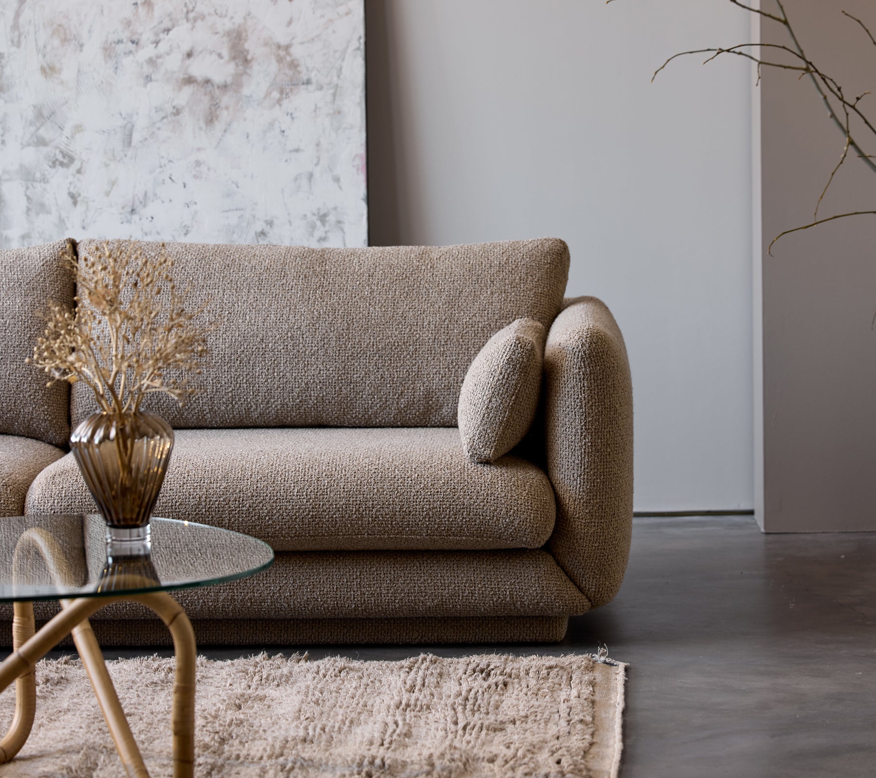 Ett taupefärgat soffbord med glasplatta står bredvid en texturerad beige soffa pyntad med en dekorativ vas och torkade blommor.