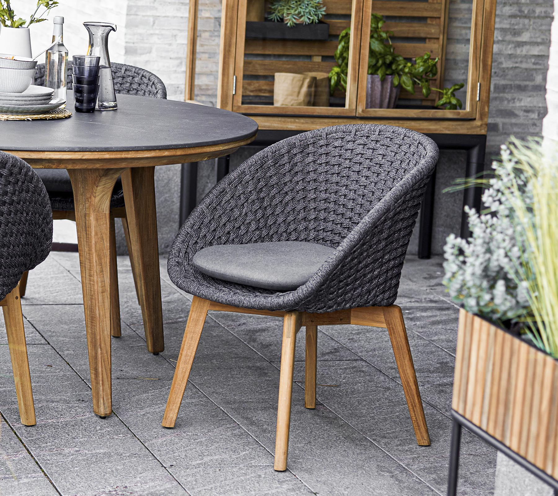 Stilfull vävd stol med en vadderad sits, i kombination med ett runt bord och omgiven av grönska i krukor. Perfekt för sociala sammankomster.