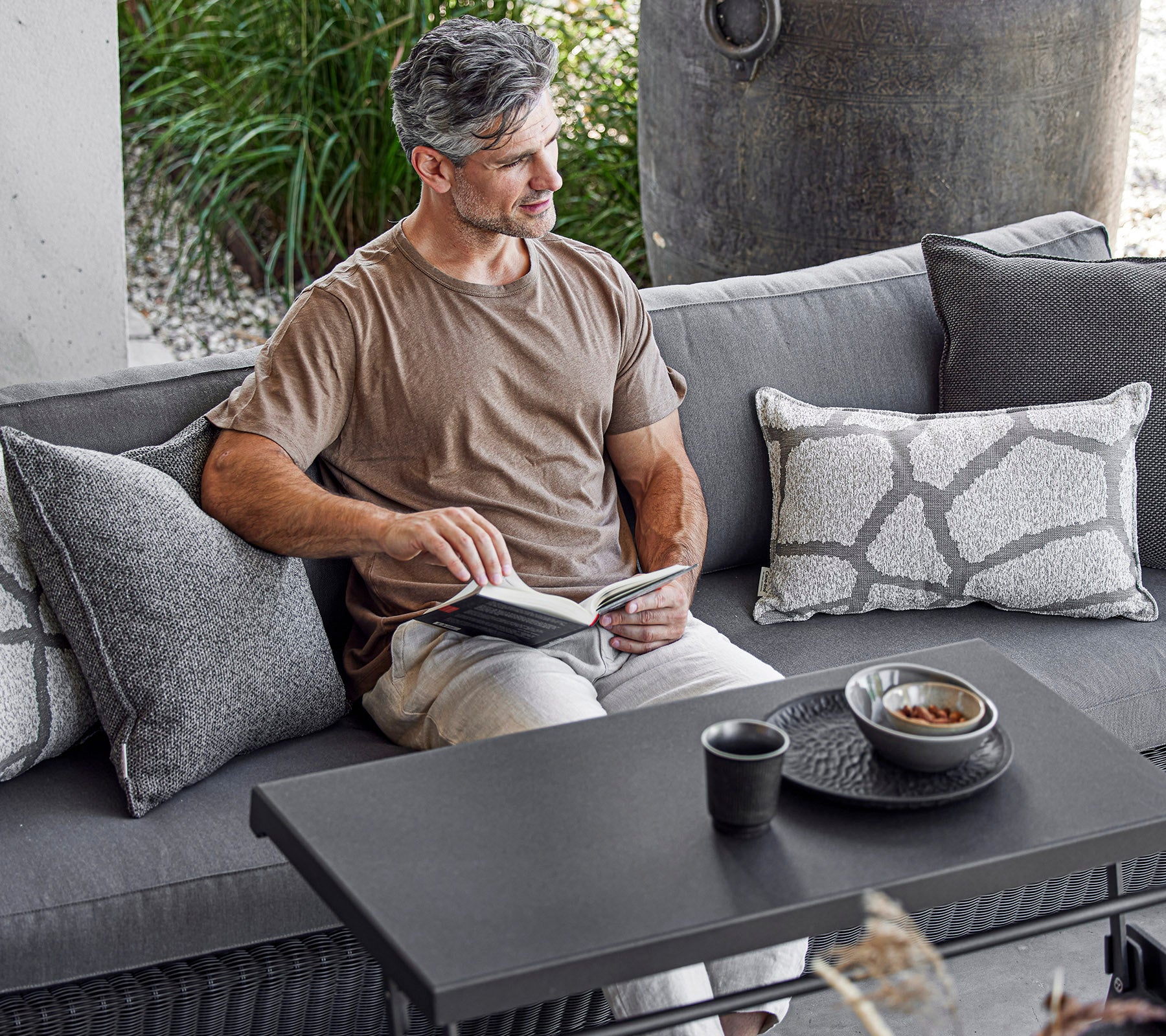 En man sitter bekvämt på en soffa och läser en bok med en kopp och en skål på ett soffbord bredvid sig, omgiven av dekorativa kuddar.