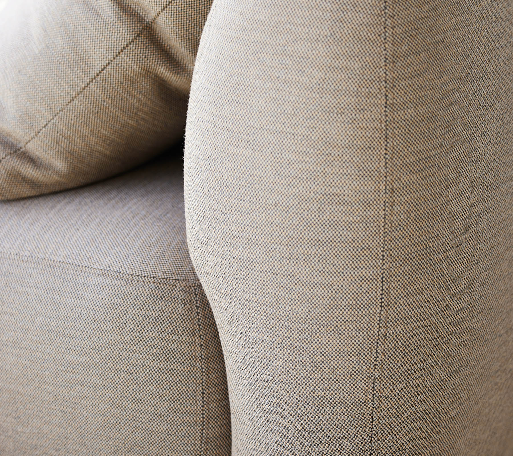 Närbild av ett texturerat beige tyg, som framhäver den böjda armstödet och de mjuka kuddarna på en möbel.