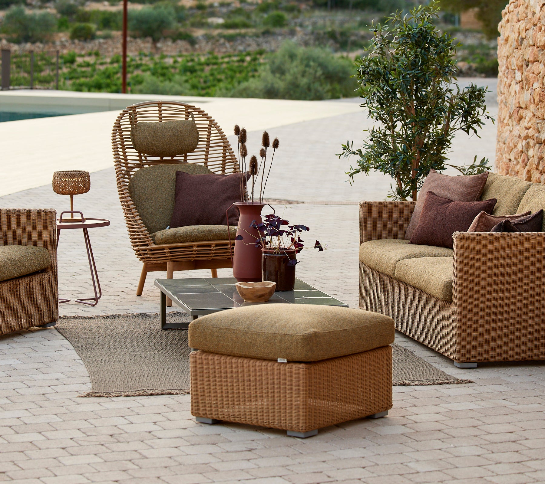 Lounge sittområde med rattanstol, beige soffa och sidobord med en lila vas.