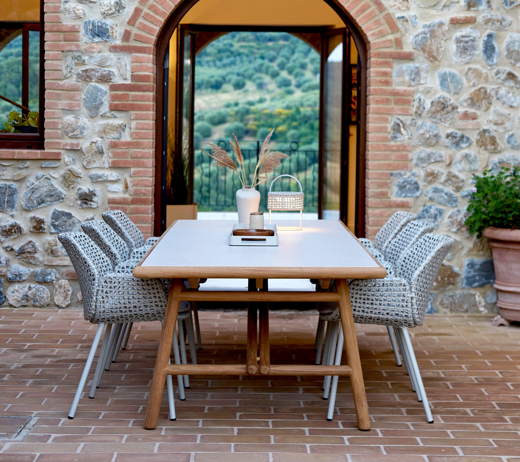 A large rectangular dining table with woven chairs, featuring a club tray centerpiece and an illusion lamp, set against a stone backdrop.