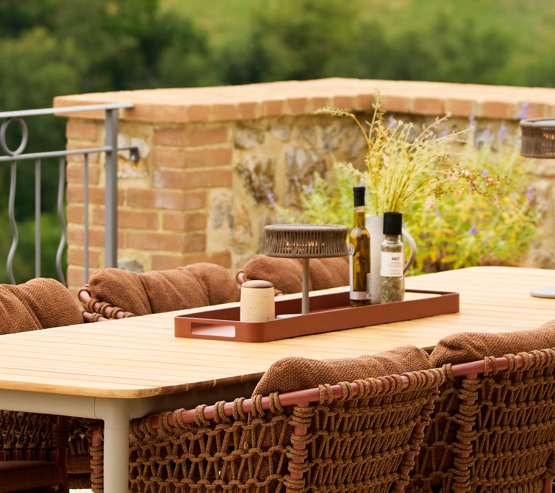 A large rectangular red tray sits on a table, featuring olive oil, a candle, and decorative elements, with warm, textured seating around it.