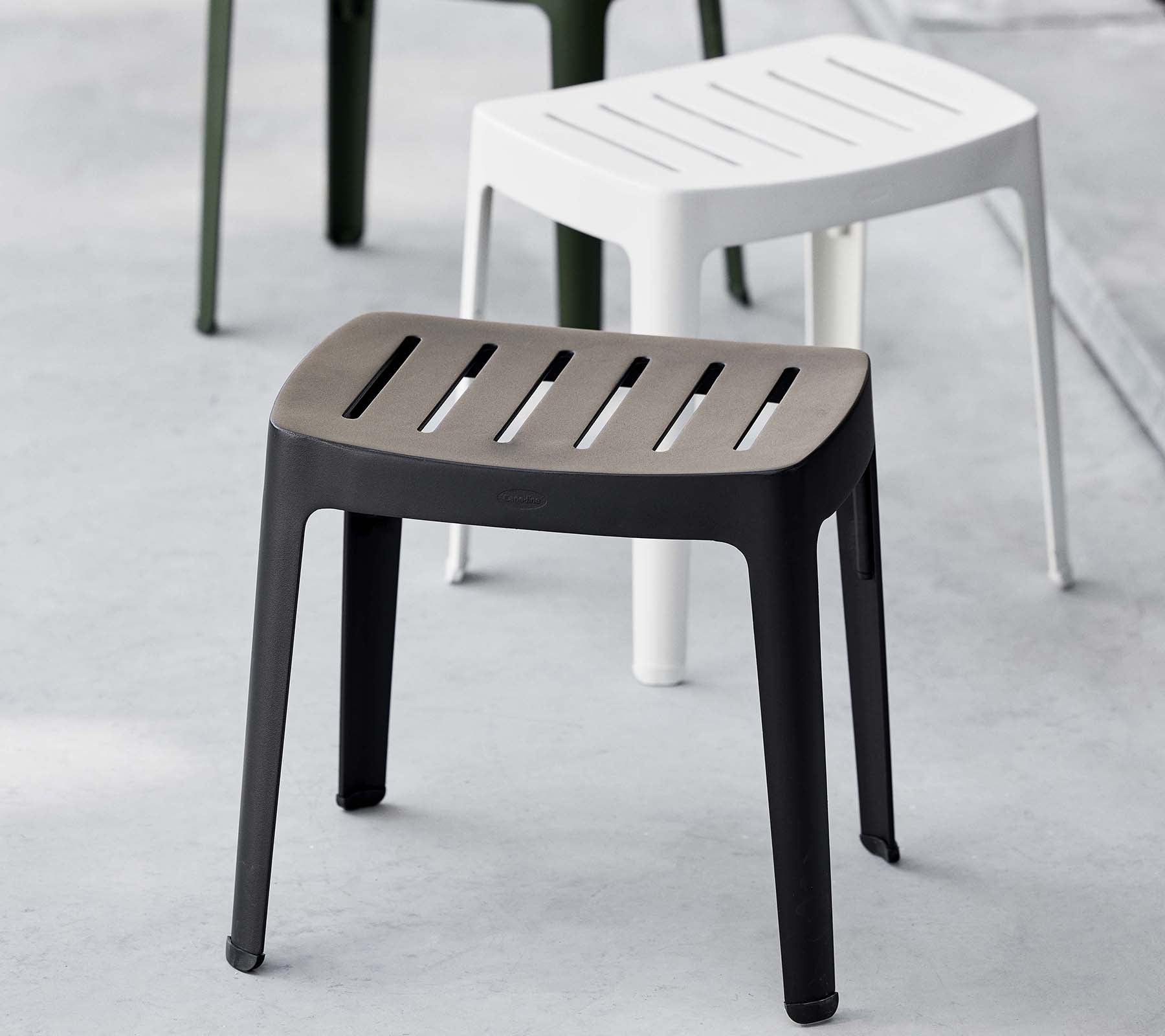 Stilren pall i svart med lamelltoppp, som har en modern design; kompletterad av en vit pall i bakgrunden.