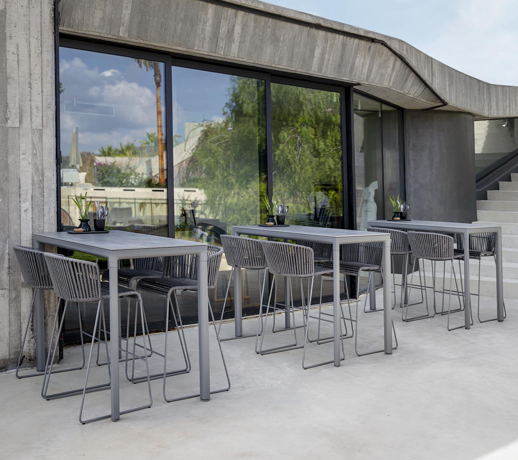 Modernt sittområde med grå bord och stolar, med stora fönster som speglar det gröna utanför. Minimalistisk designestetik.