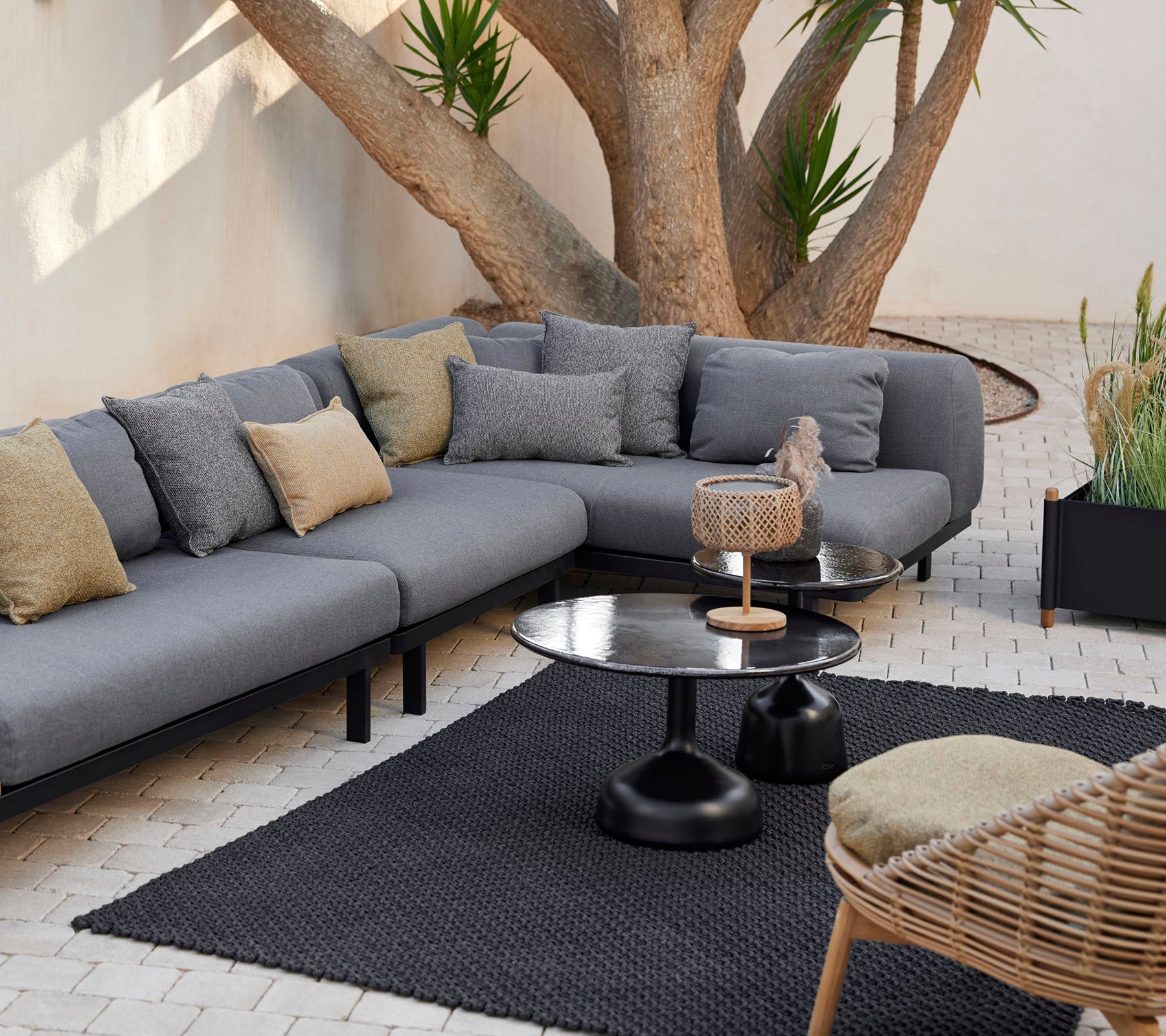 Loungeområde med en grå modulsoffa med dekorativa kuddar, ett stilrent soffbord och en texturerad matta omgiven av grönt.