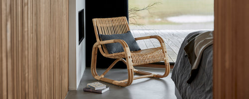 Vävd stol med en bekväm kudde, placerad bredvid en säng, som visar en modern och stilren design i en lugn miljö.