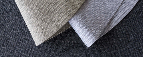 Två texturerade tygstycken i neutrala toner, placerade diagonalt på en cirkulär mörk bakgrund, som visar upp sina subtila mönster.