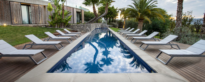 En lugn poolscen med en rad vita solsängar vid sidan av ett stilla vatten, omgiven av frodig grönska och palmer.