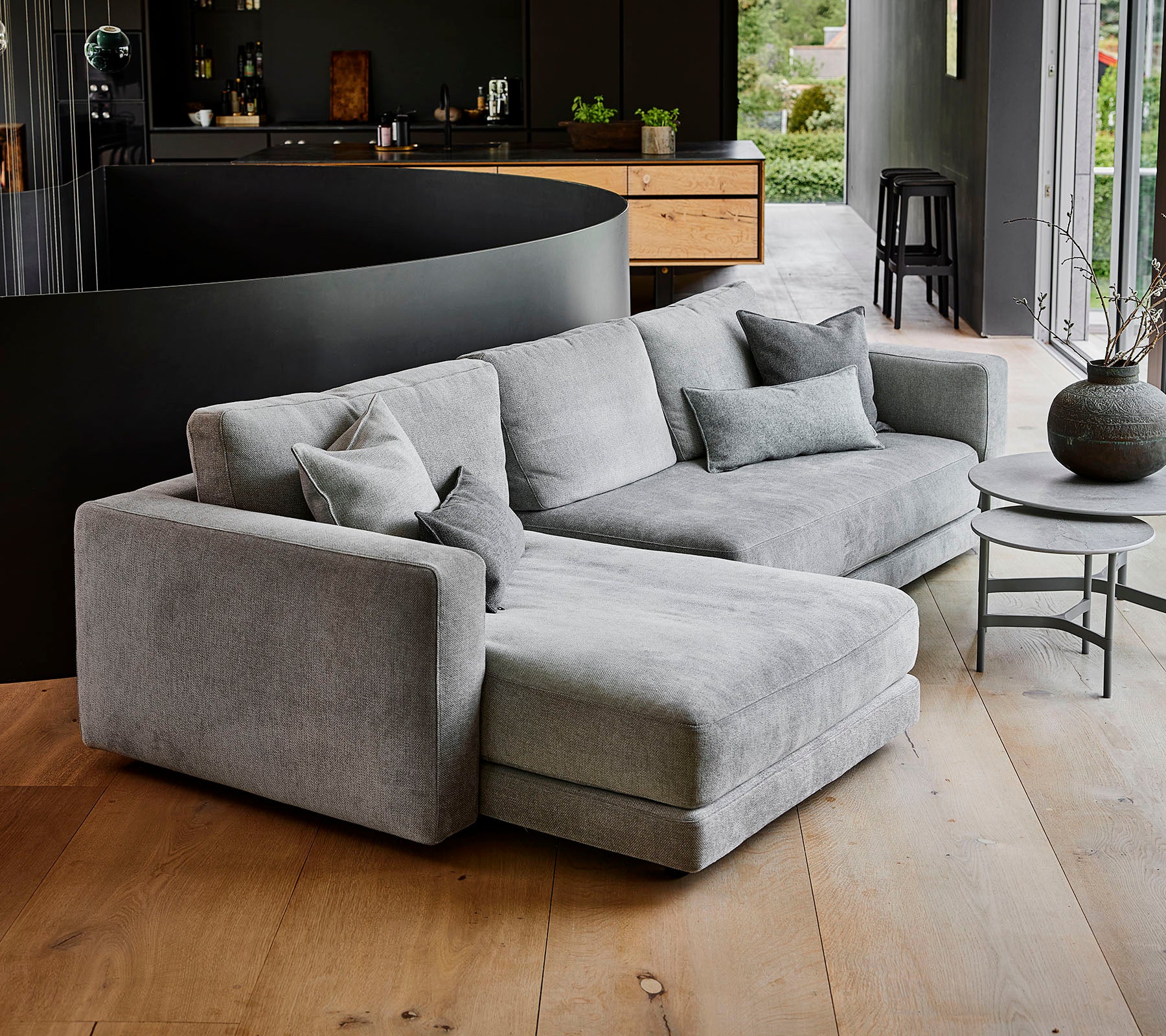 Modern grå hörnsoffa med flera kuddar, tillsammans med ett runt soffbord och en dekorativ vas. Enkelt, elegant design.
