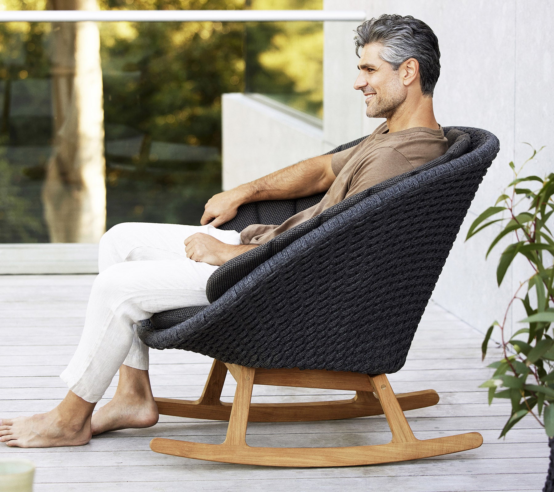 En man sitter avslappnad i en stilren, modern gungstol med en vävd design och en bas av naturligt trä, omgiven av grönska.