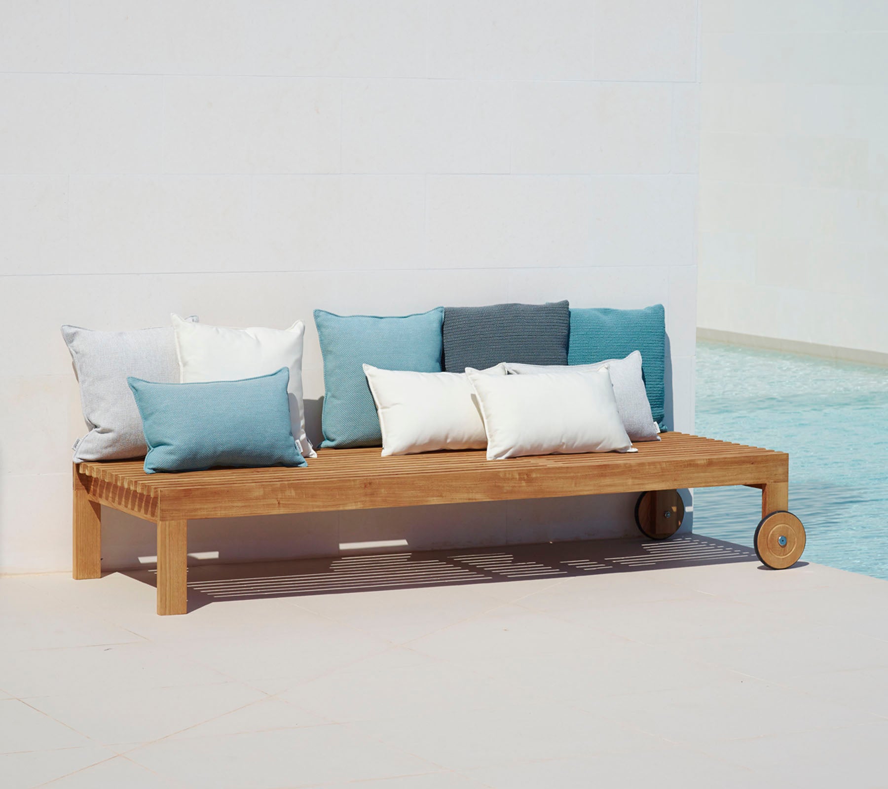 Trä loungesoffa med enkel design, med kuddar i blå och vita nyanser, placerad bredvid en pool.