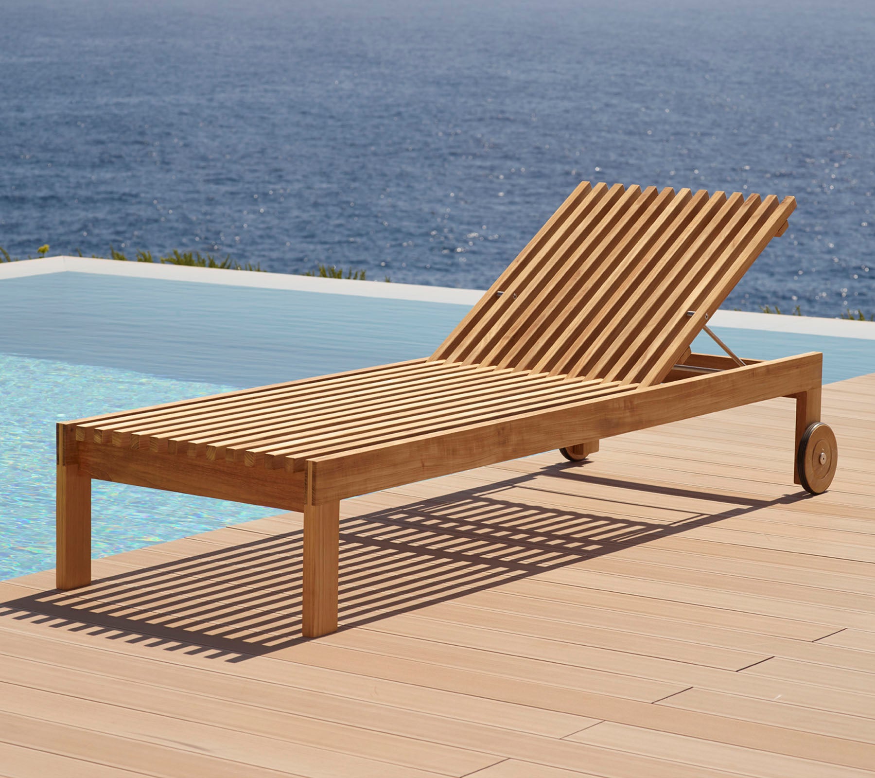 Trä-loungestol med slattedesign, utrustad med hjul och placerad vid en swimmingpool med vattenutsikt i bakgrunden.