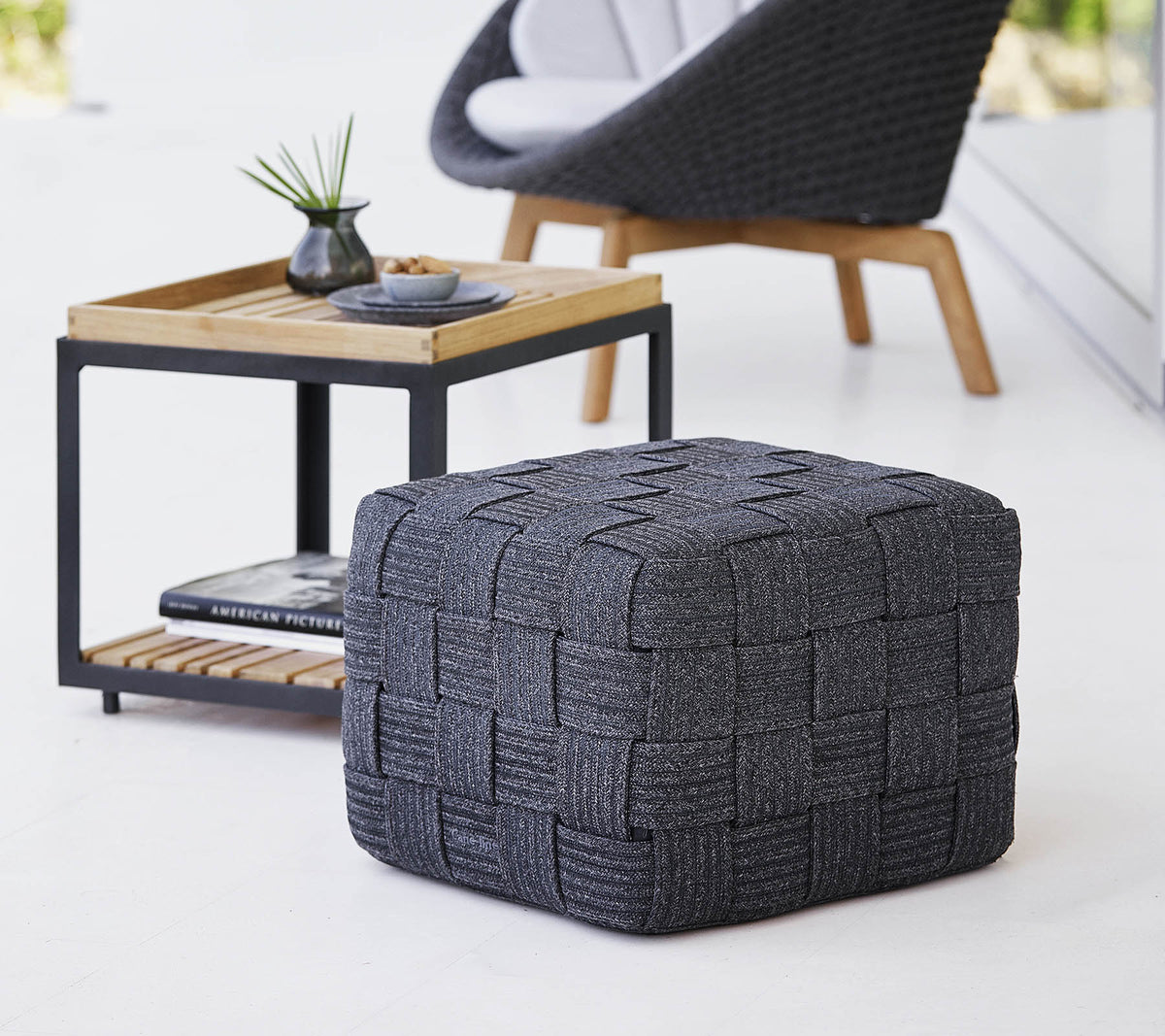 En texturerad grå vävd pouf placerad nära ett träsidebord med minimalistisk design, som har små dekorationsföremål.