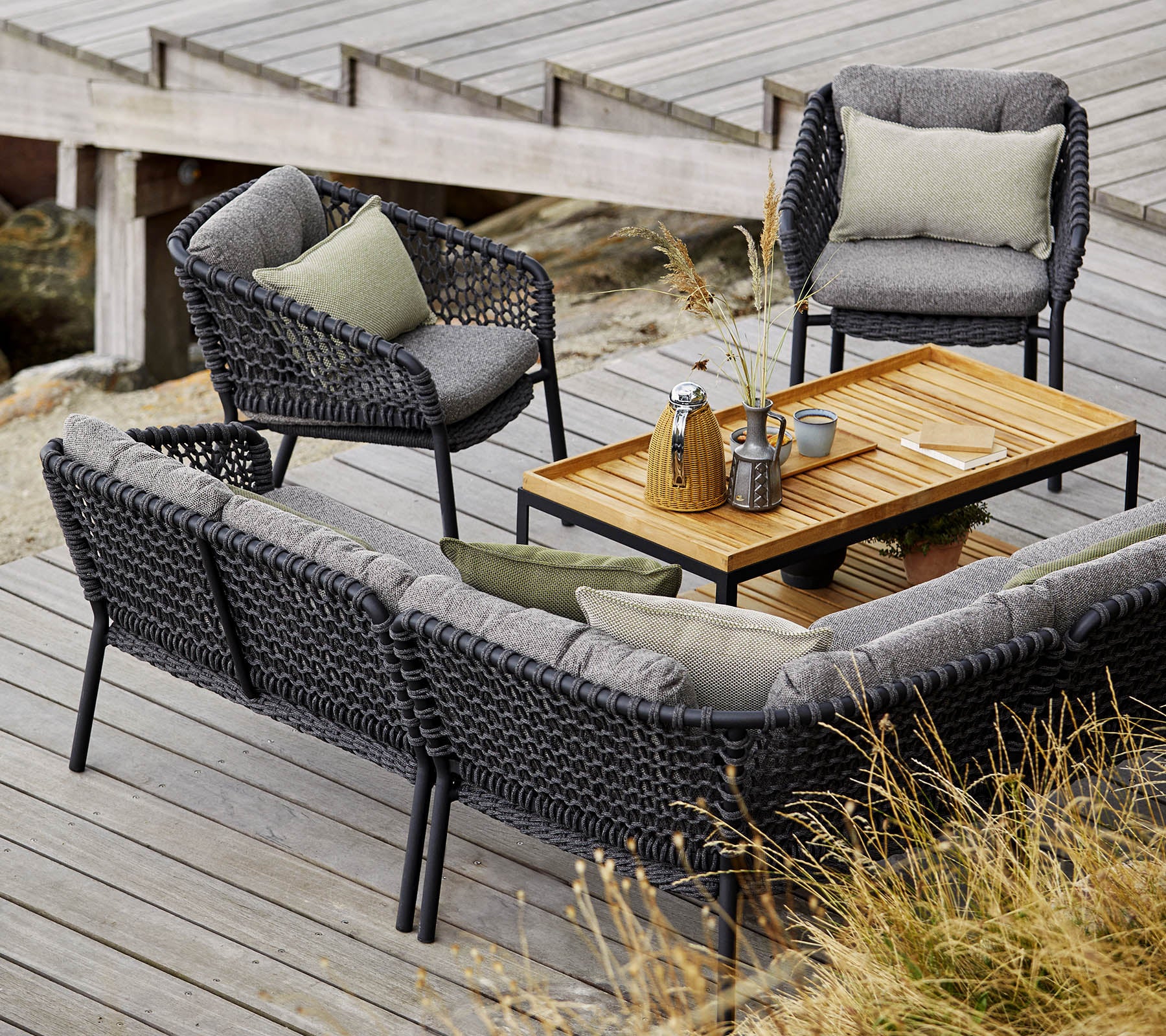 Stilfullt utemöbelset med en mysig soffa, två fåtöljer och ett träkaffebord, präglat av mjuka kuddar och dekorativa föremål.