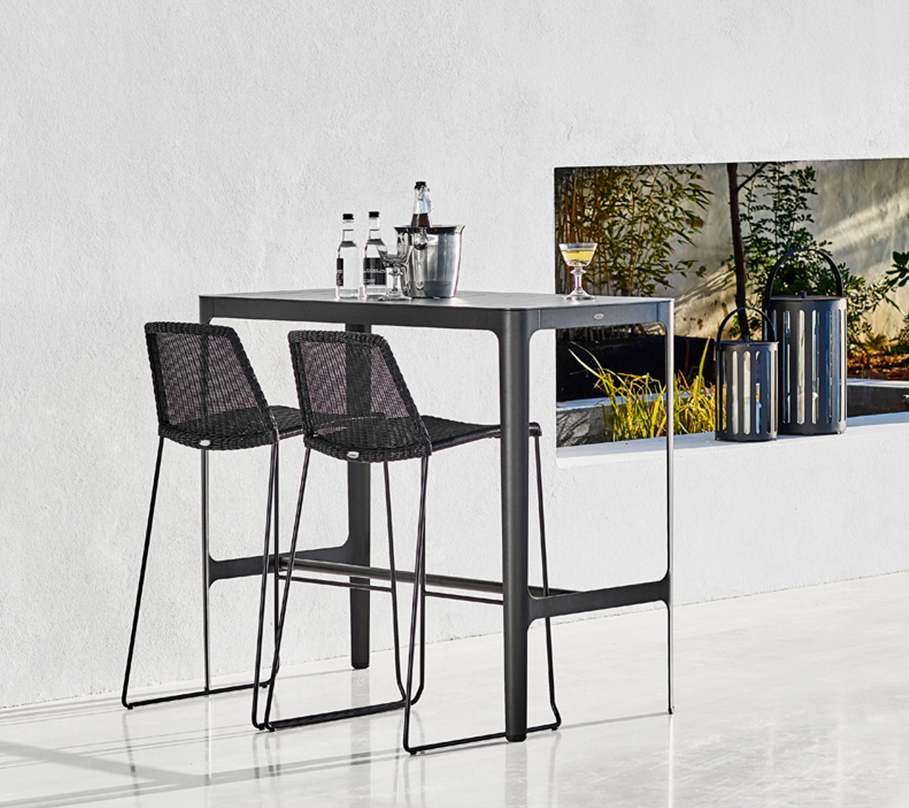 Elegant bardisk med två svarta, vävda barstolar, ackompanjerad av glas och en stilren hink, mot en minimalistisk bakgrund.