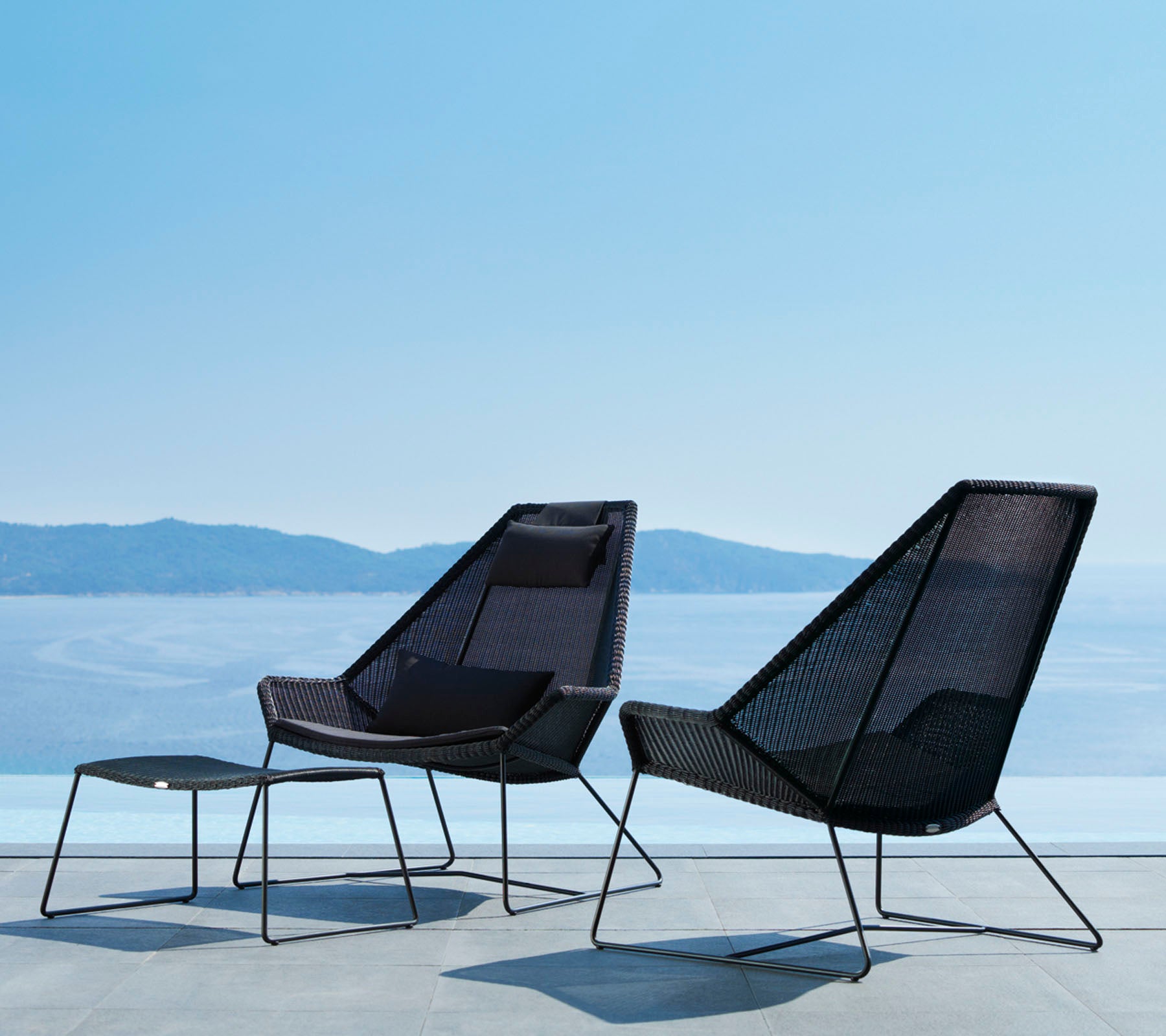 Två stiliga, moderna stolar med eleganta linjer och öppet mesh-design, tillsammans med en matchande fotpall, med utsikt över en lugn vattenutsikt.