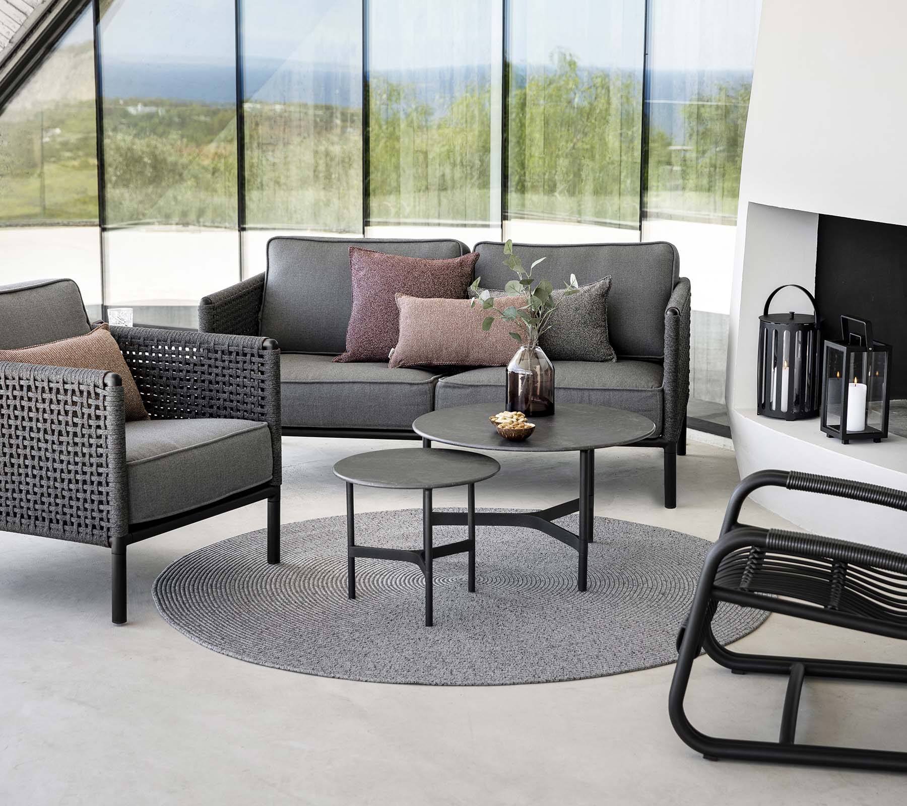 Stilfullt sittområde med en grå soffa med kuddar, runt soffbord och accentstolar på en rund matta. Naturligt ljus förbättrar atmosfären.