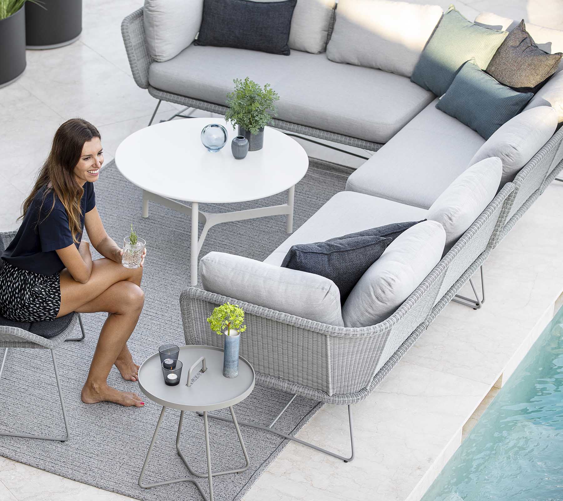 En stilren utomhus sittgrupp med en soffa, prydkuddar och ett litet bord, kompletterat med växter och en pool i närheten.