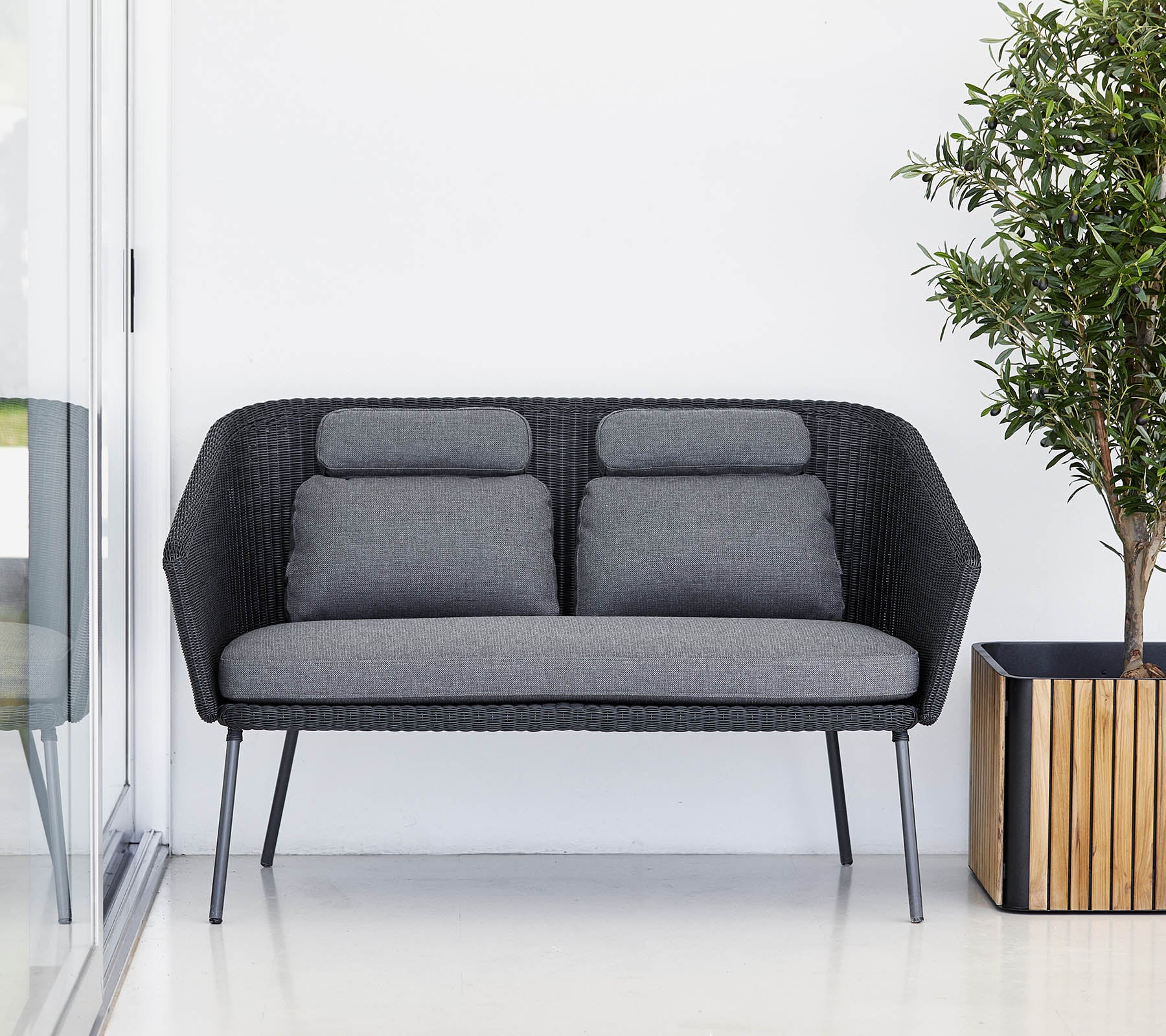 Stilsoffan i svart med grå kuddar, kompletterad med en krukväxt i en träkruka. Modernt och minimalistiskt design.