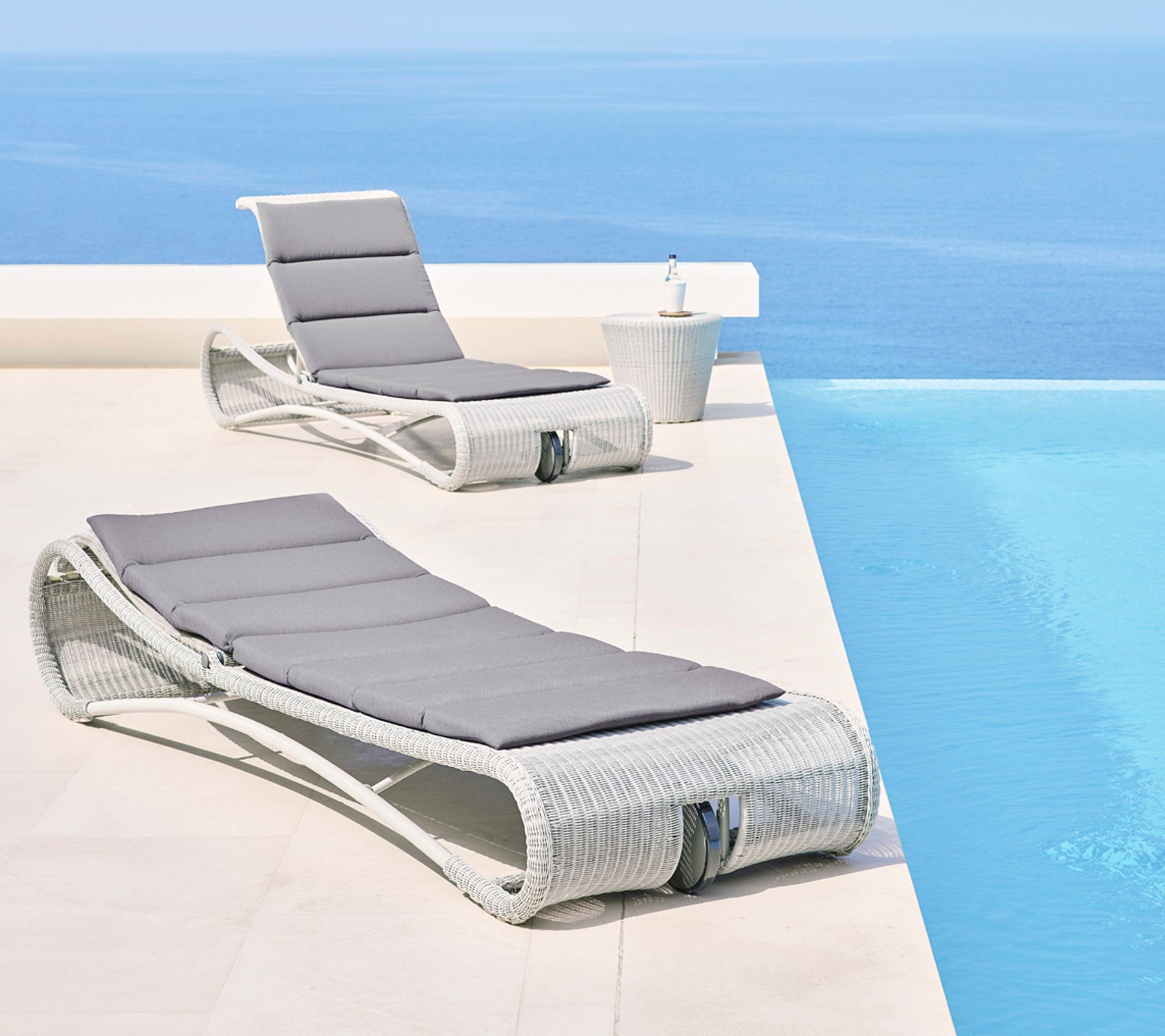 Två stiliga loungestolar med grå kuddar bredvid en lugn blå infinitypool, vilket skapar en avkopplande utomhusmiljö.
