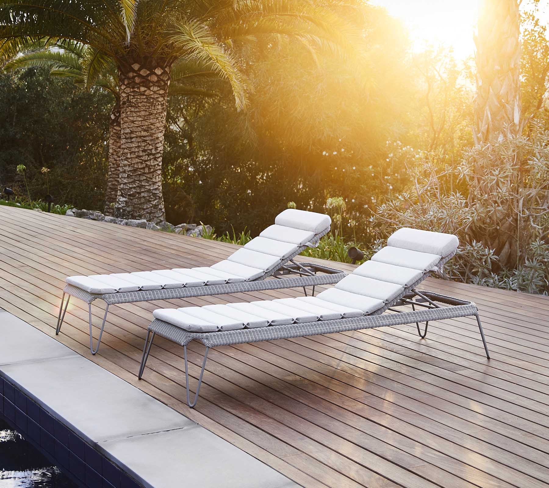 Två stiliga loungefåtöljer med vita kuddar, placerade vid en pool och omgivna av frodig grönska som njuter av det varma solskenet.