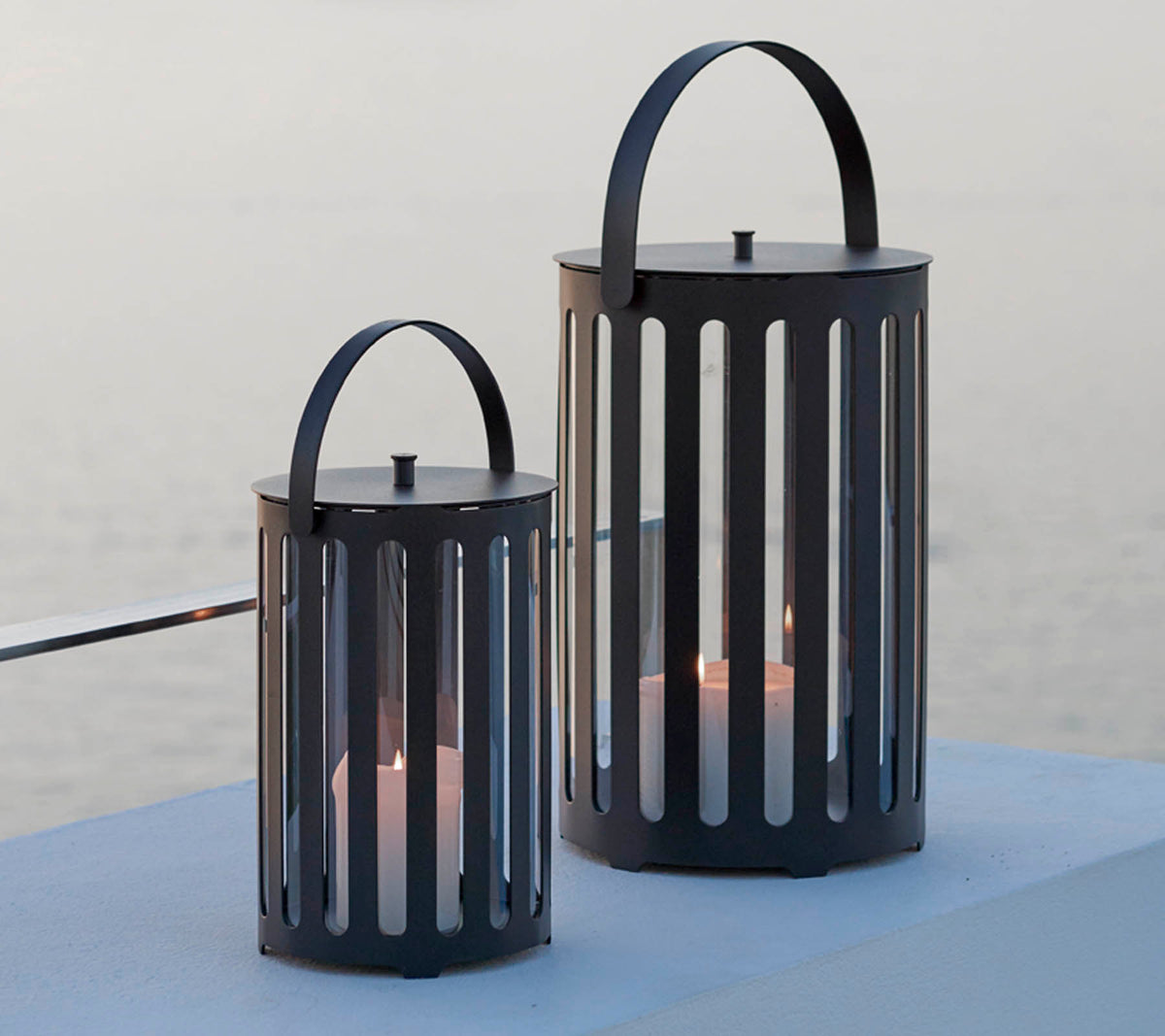 Två stiliga metallampor med vertikala springor, med handtag och ljus inuti, mot en lugn bakgrund.