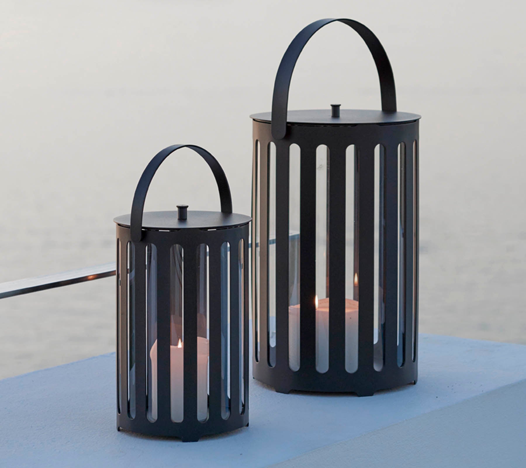 Två stiliga metallampor med vertikala springor, med handtag och ljus inuti, mot en lugn bakgrund.