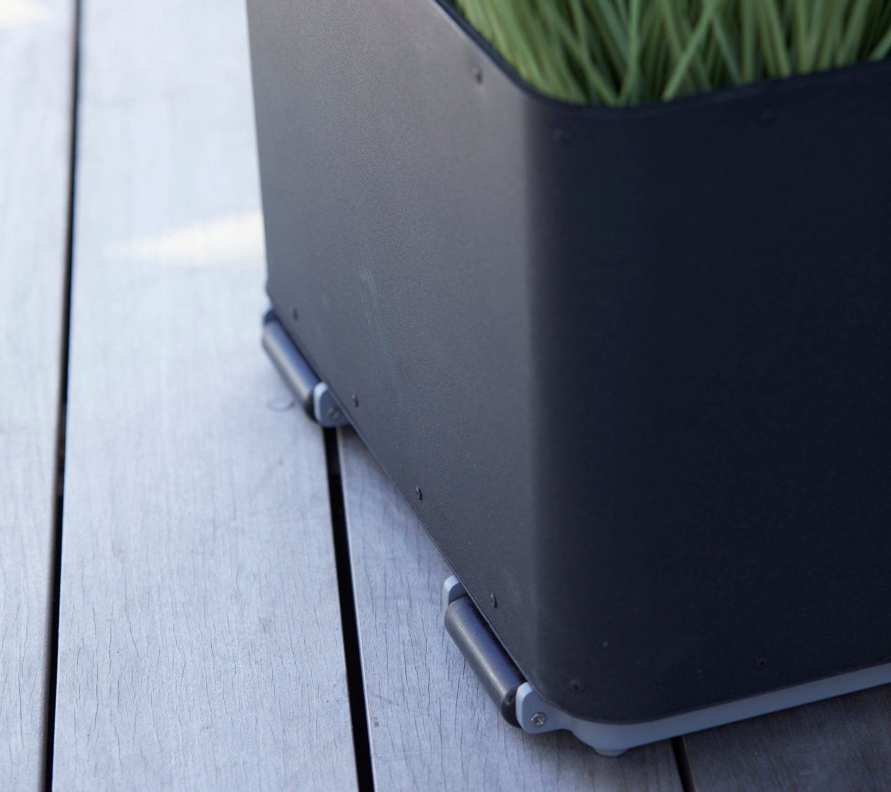 Svart planterlåda med en elegant design, som har grön gräs och silverkopplingar som sitter på en träyta.