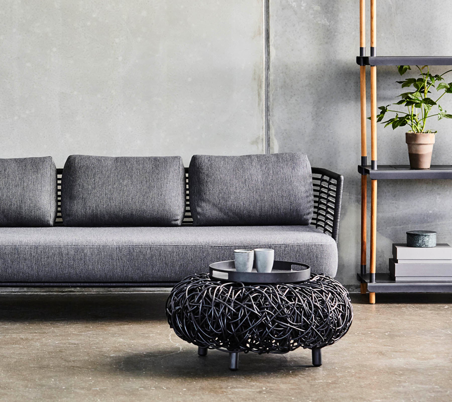 En modern grå soffa med kuddar, tillsammans med ett runt svart vävt soffbord och en minimalistisk hylla som visar växter och dekoration.